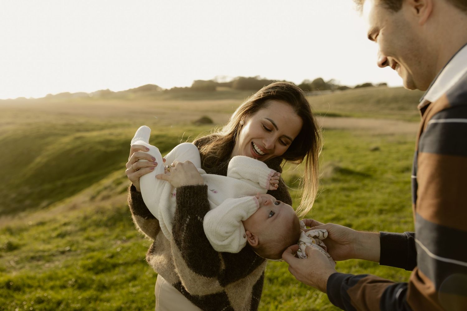 Parents joyfully playing with their baby outdoors in golden sunlight on a green grassy hill.
