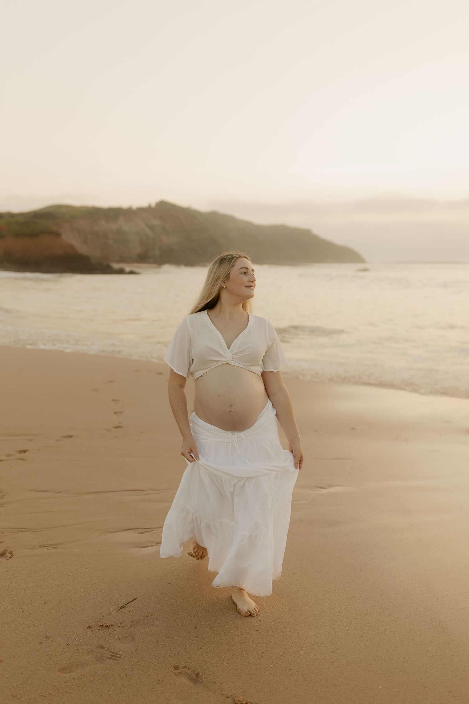 A dreamy beach photoshoot at sunset shows a figure in a flowing white dress dancing on golden sand.