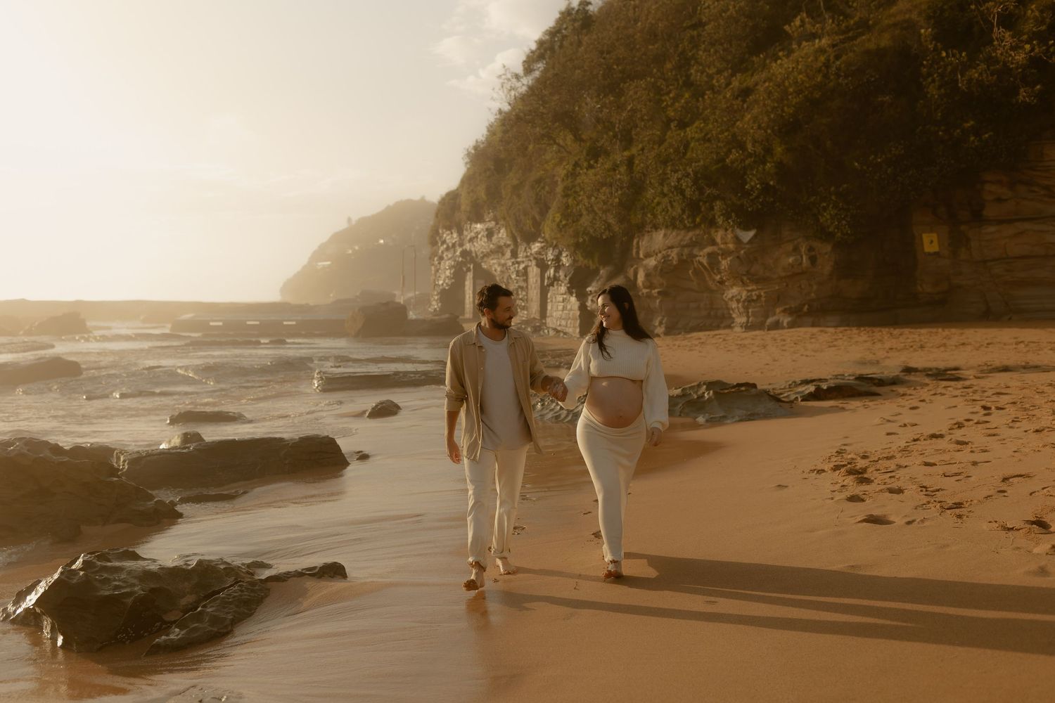 A couple walks along a misty beach shoreline with rock formations and cliffs visible in the warm evening light.
