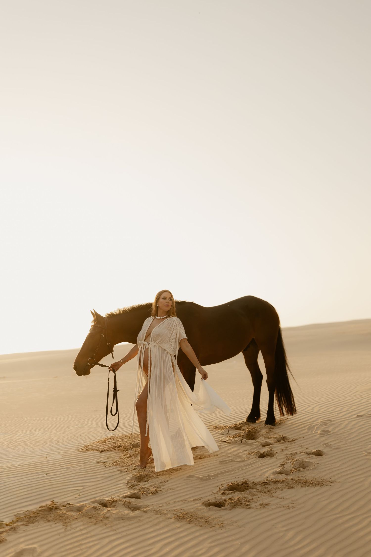 Person in flowing white dress stands beside majestic horse on desert sand dunes at golden hour.