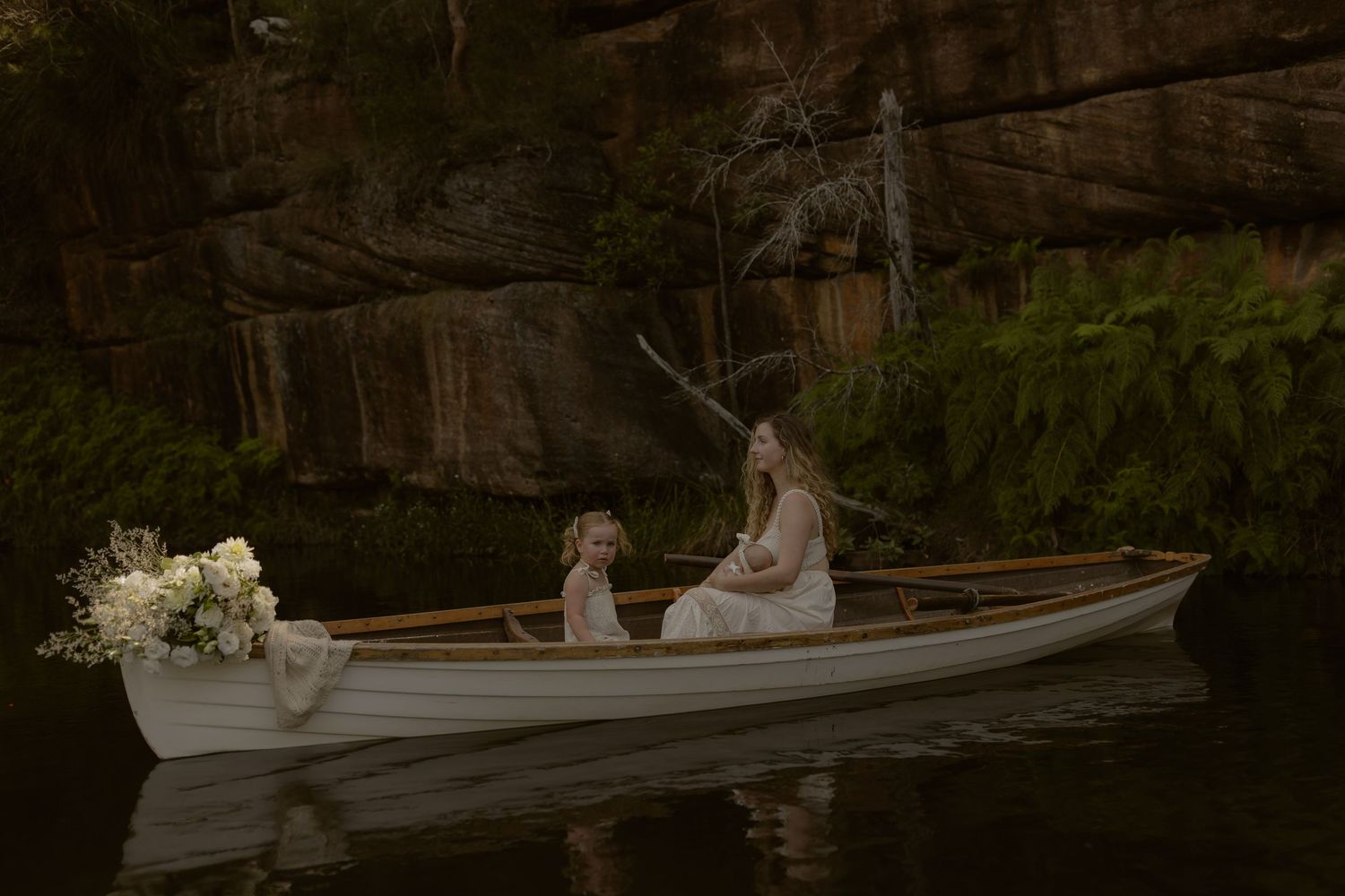 A white wooden rowboat sits on calm water surrounded by trees and greenery at sunset.