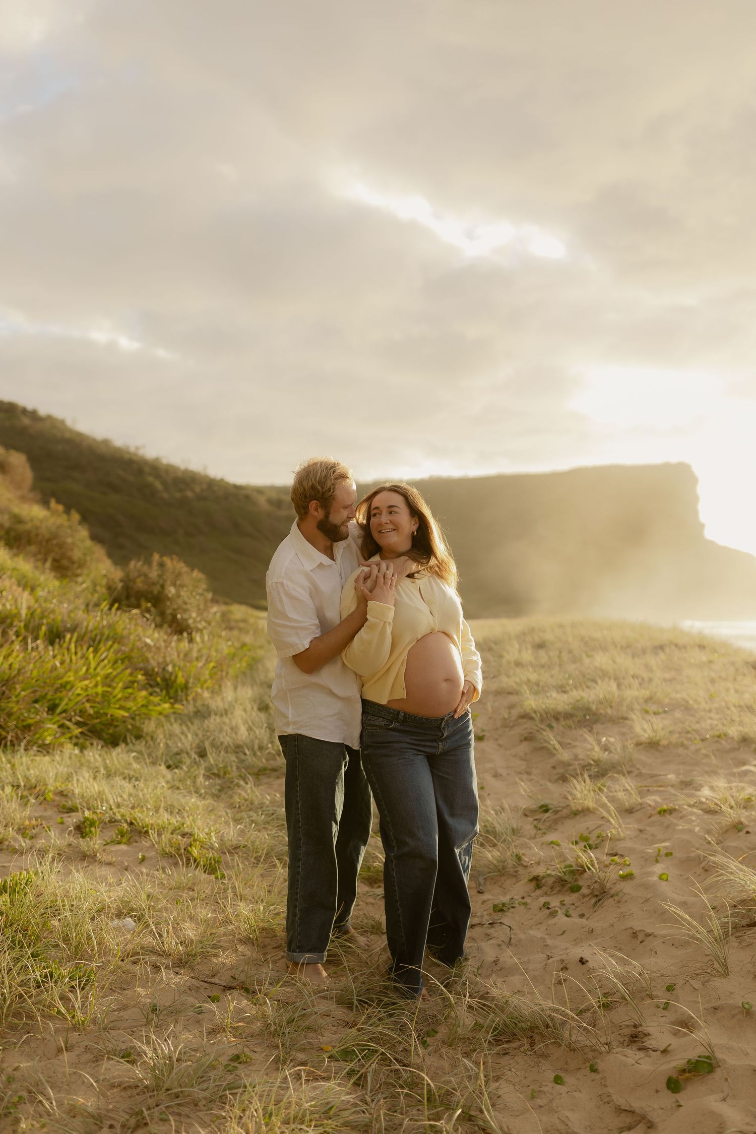 Romantic moment shared between two people on a grassy coastal hillside at sunset.