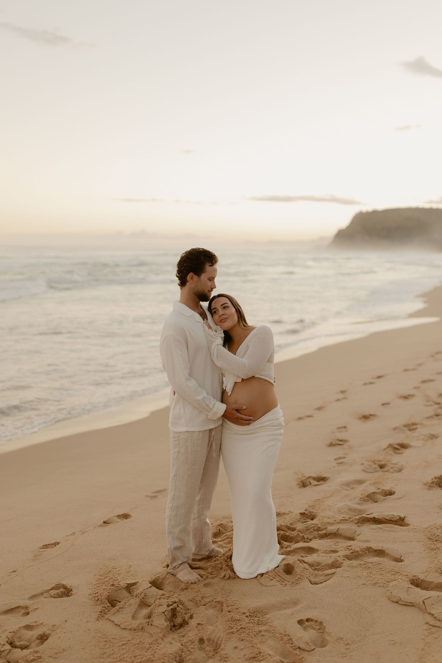 A romantic beach photoshoot with footprints in the sand as a couple embraces near the ocean.