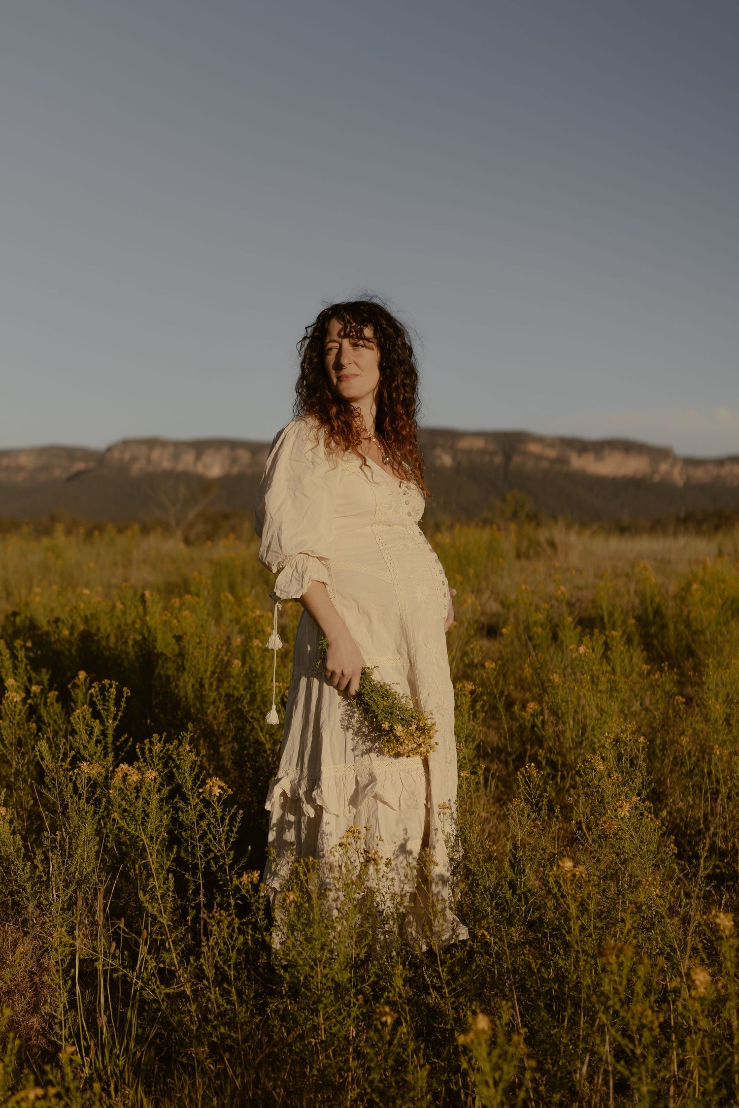 A figure in a flowing white dress stands in a golden meadow at sunset with mountains in the background.
