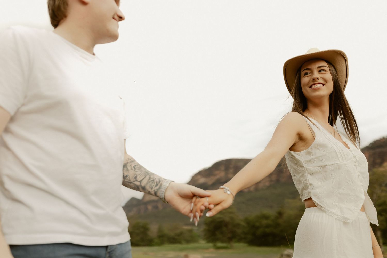 Couple in white outfits share intimate moments during outdoor photo session at sunset with mountain backdrop.
