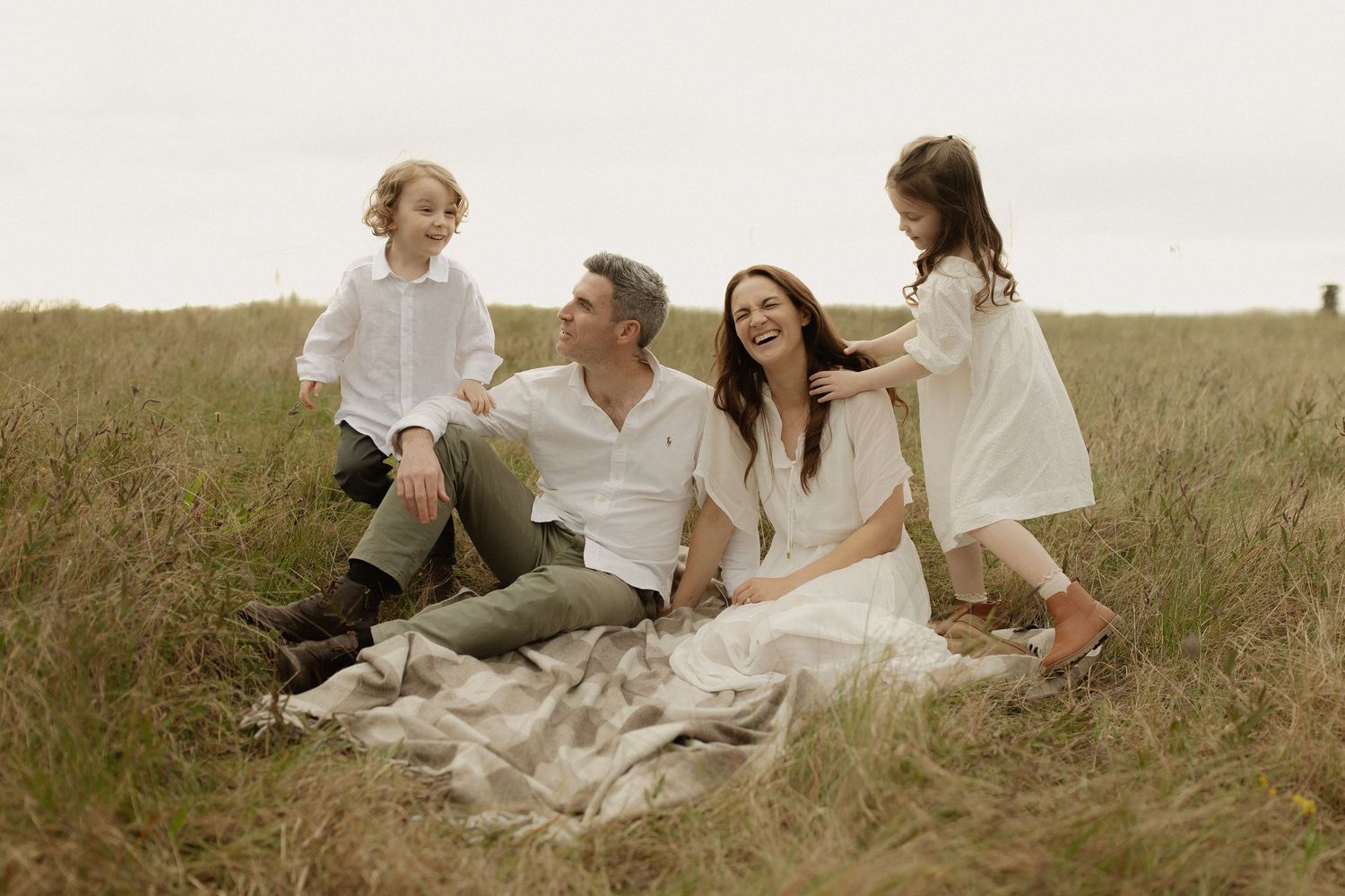 A joyful group sits together on a blanket in a golden grassy field during a casual outdoor photo session.