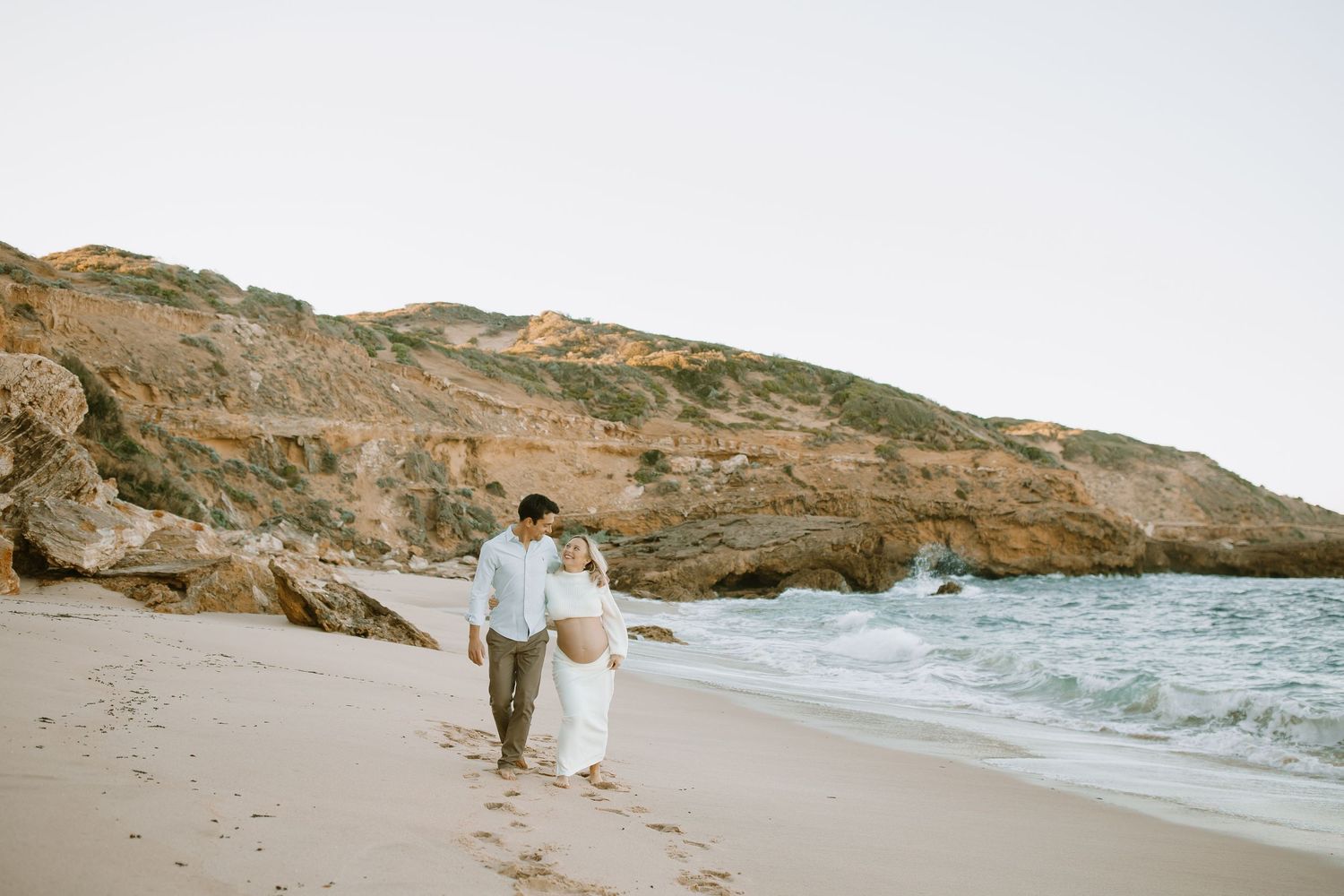 A couple walks together on a scenic coastal beach surrounded by rugged cliffs at sunset.