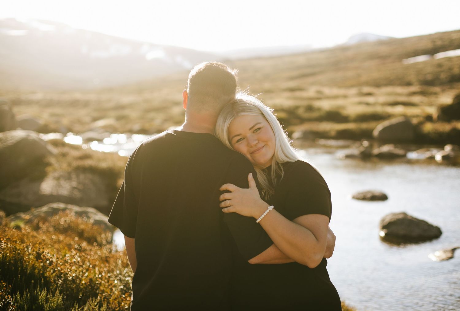 A couple embraces beside a scenic mountain lake during golden hour with rocks and hills in the background.