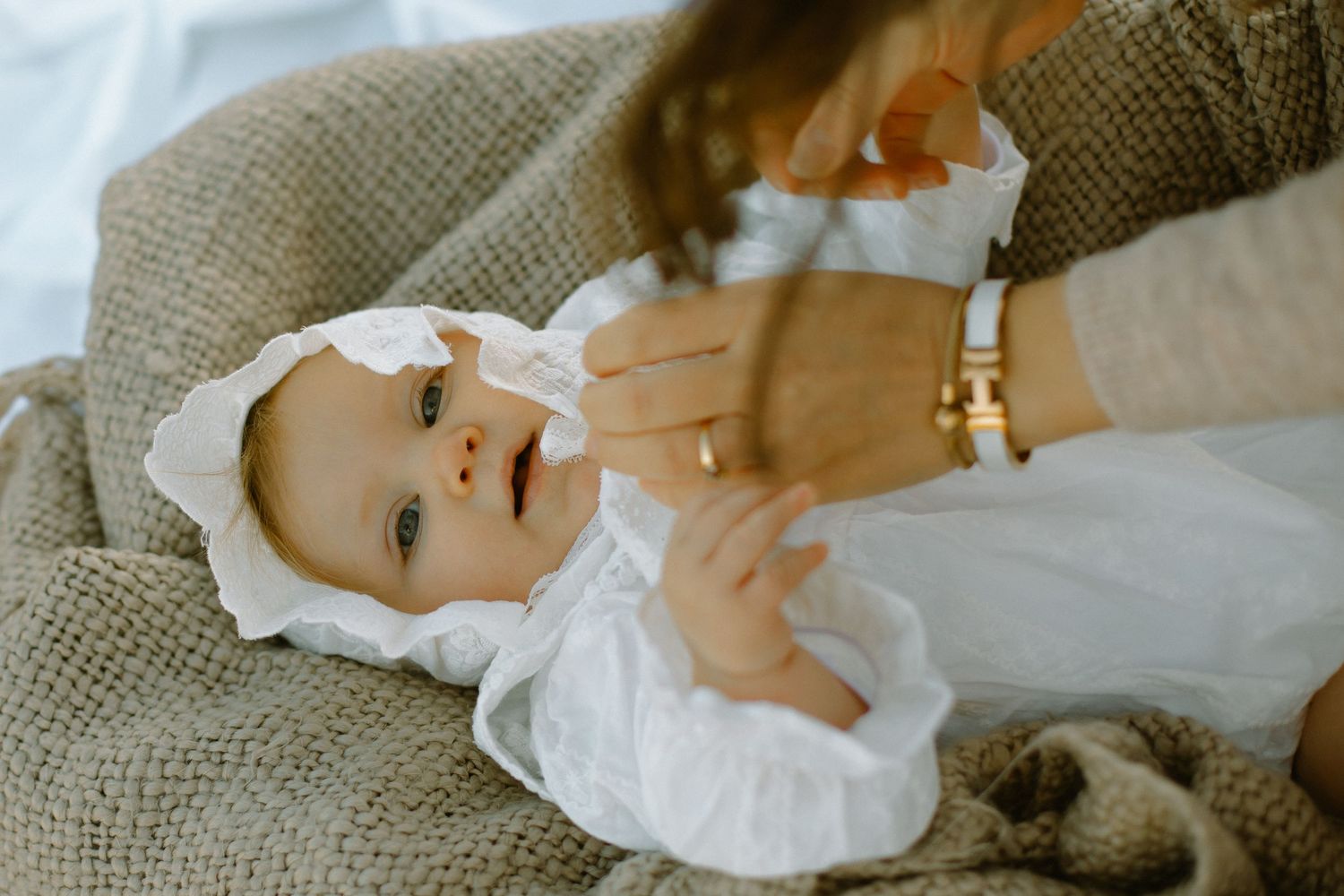 Baby in white bonnet and dress lying on textured blanket.