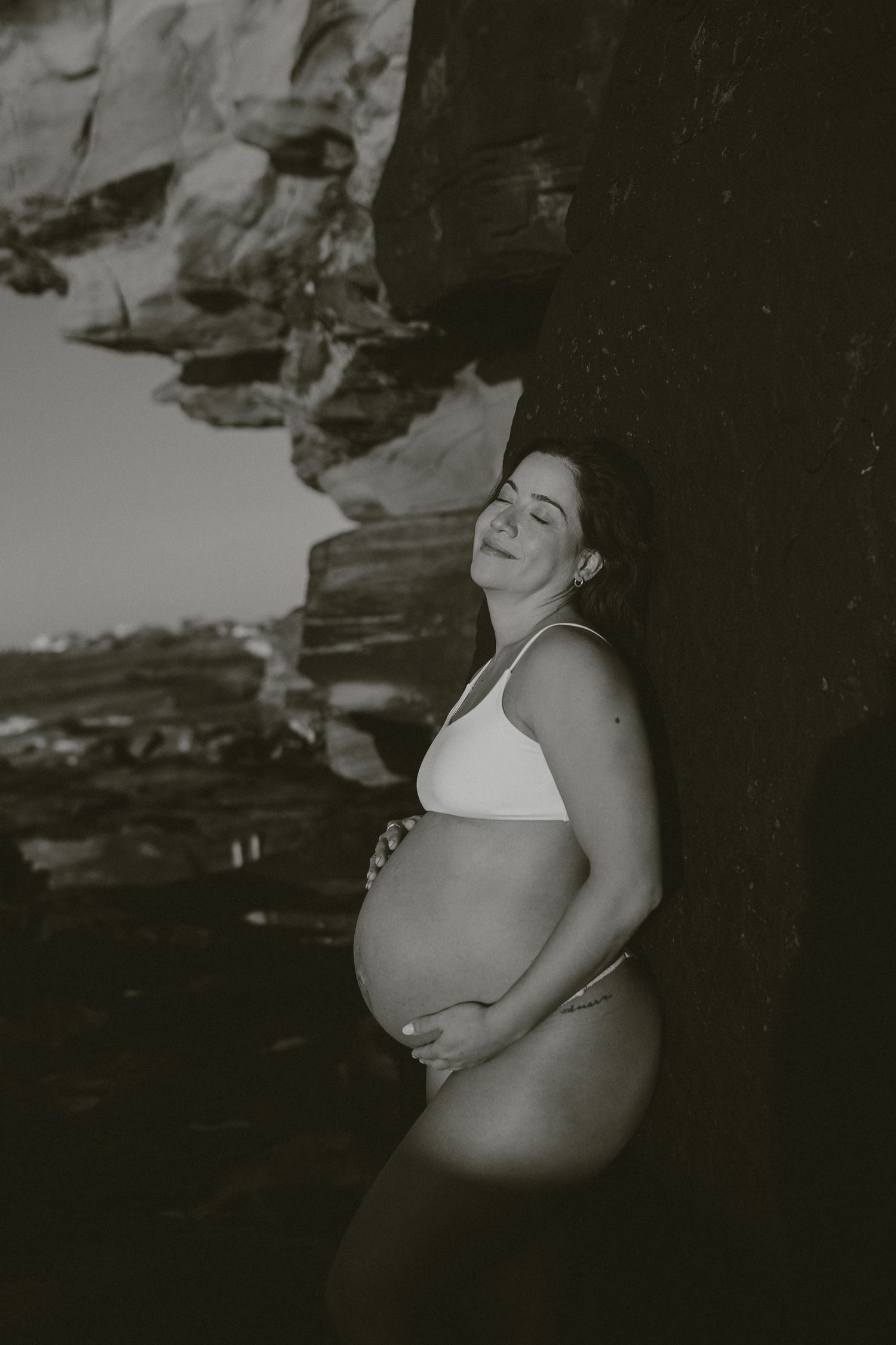 Expectant mother in white bikini poses against rocky cliffside in black and white photo.
