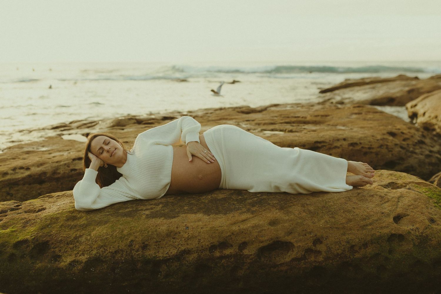 A person in white attire lying peacefully on a rocky seaside cliff during golden hour as seabirds fly in the distance.