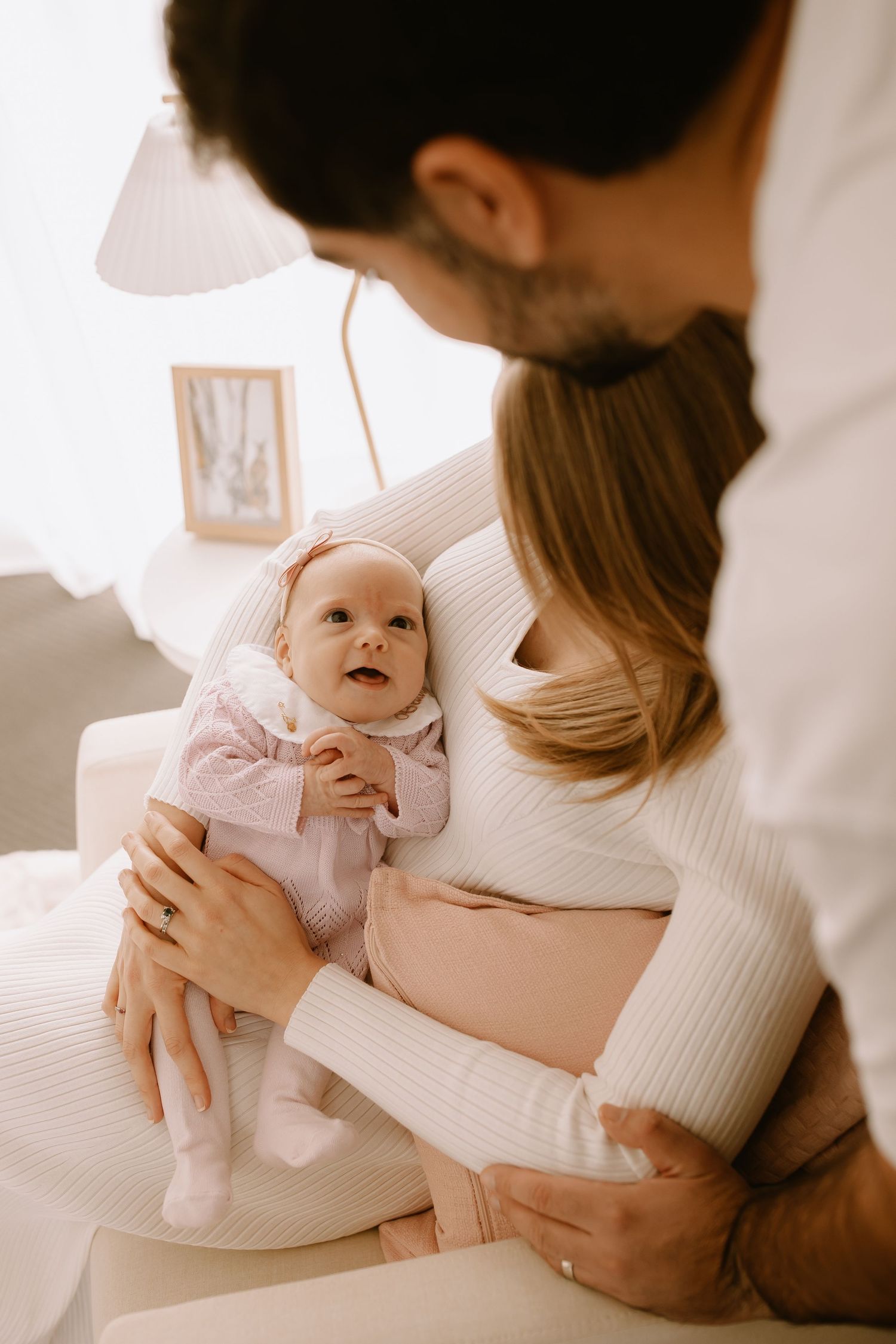 Parents in white sweaters lovingly hold their smiling baby in pink sleepwear during an intimate family moment.