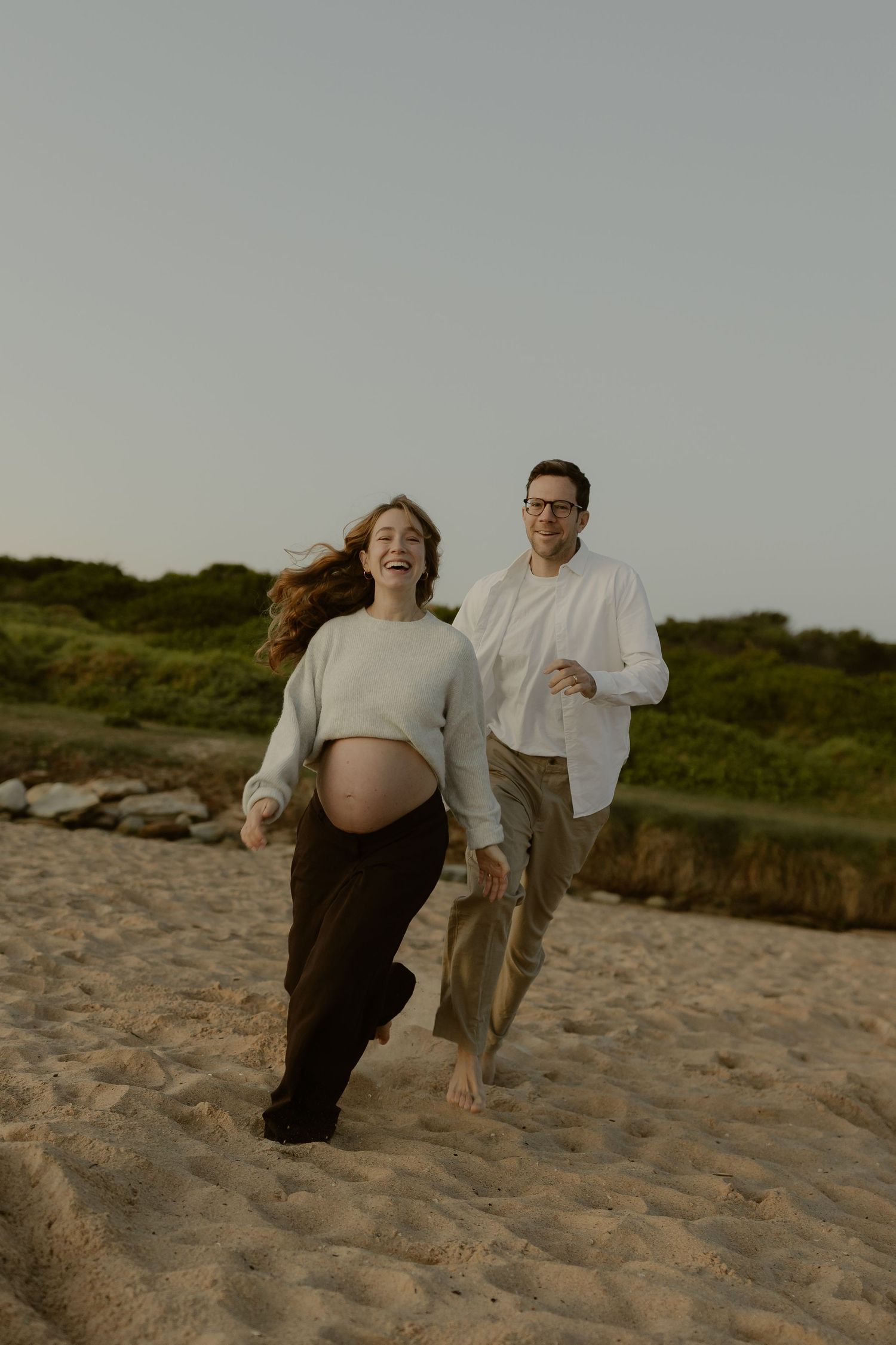 Laughing pregnant woman runs barefoot on sandy beach with partner during golden hour maternity photoshoot.