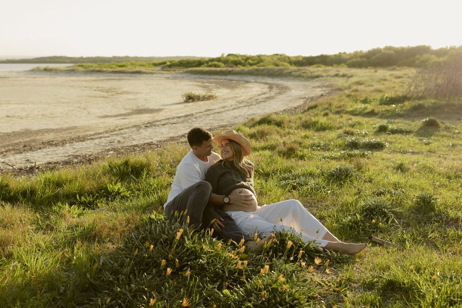 Couple sitting together on green coastal grass near a sandy beach at golden hour sunset.