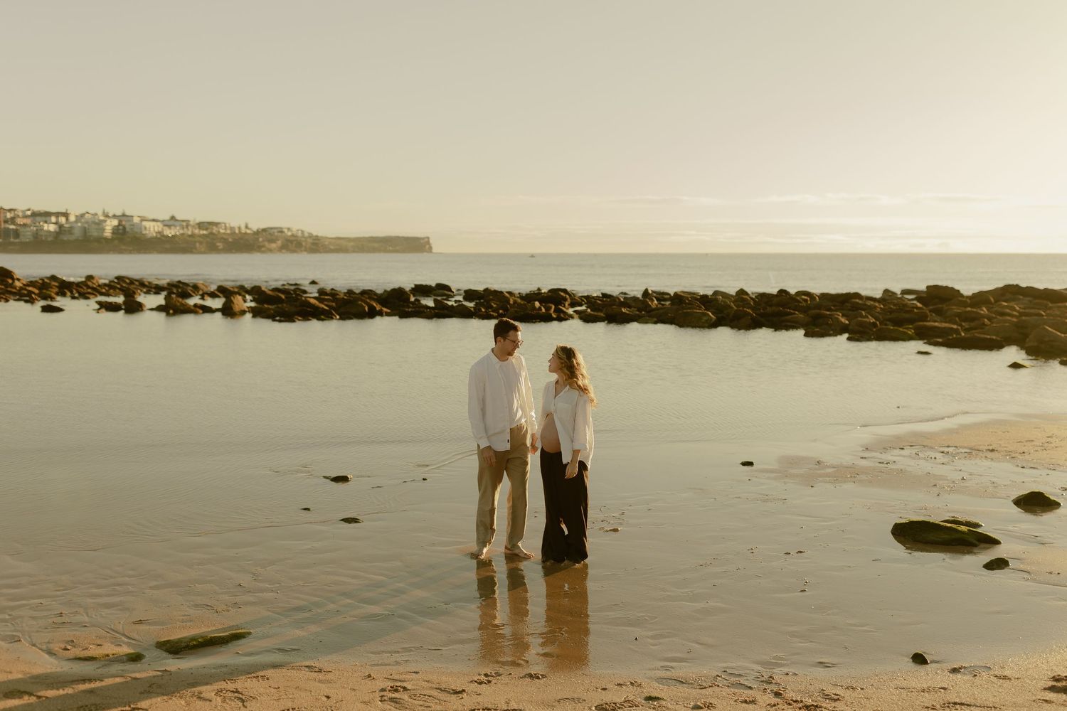 Couple in white tops holds hands facing each other on sandy beach with rocky coastline at sunset.