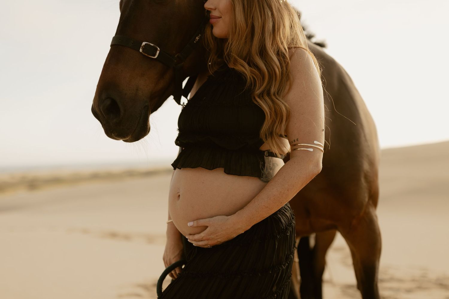 Close-up of pregnant woman cradling baby bump beside a brown horse on sandy dunes.