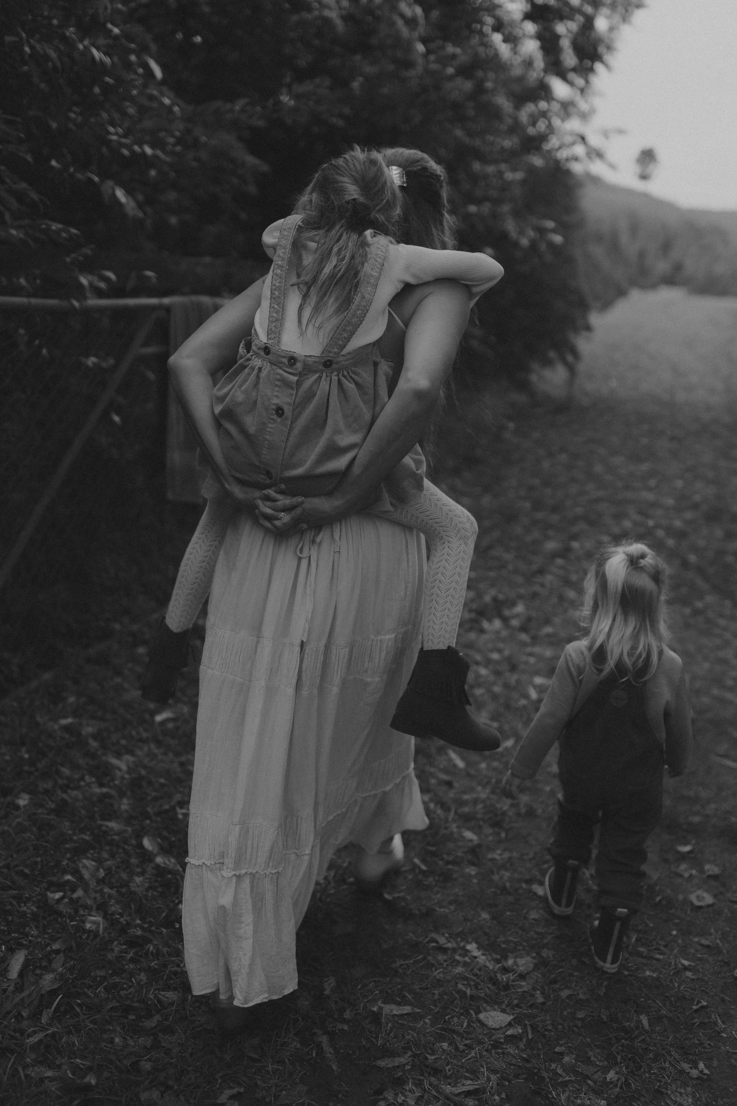 A mother in a white dress carries a child while holding another childs hand on a rural path.