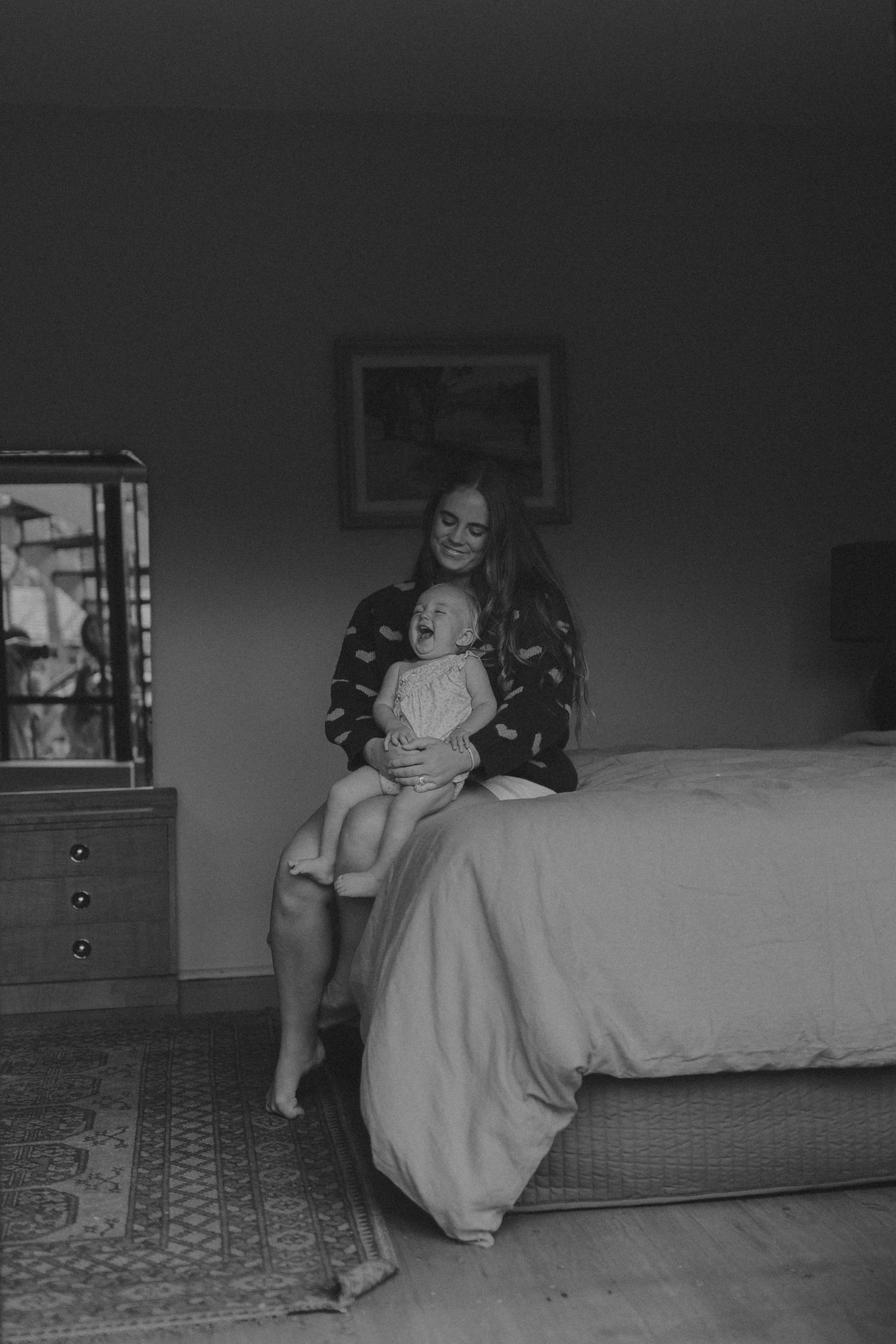 Black and white photo of a smiling mother sitting on a bed holding her laughing toddler in a cozy bedroom.