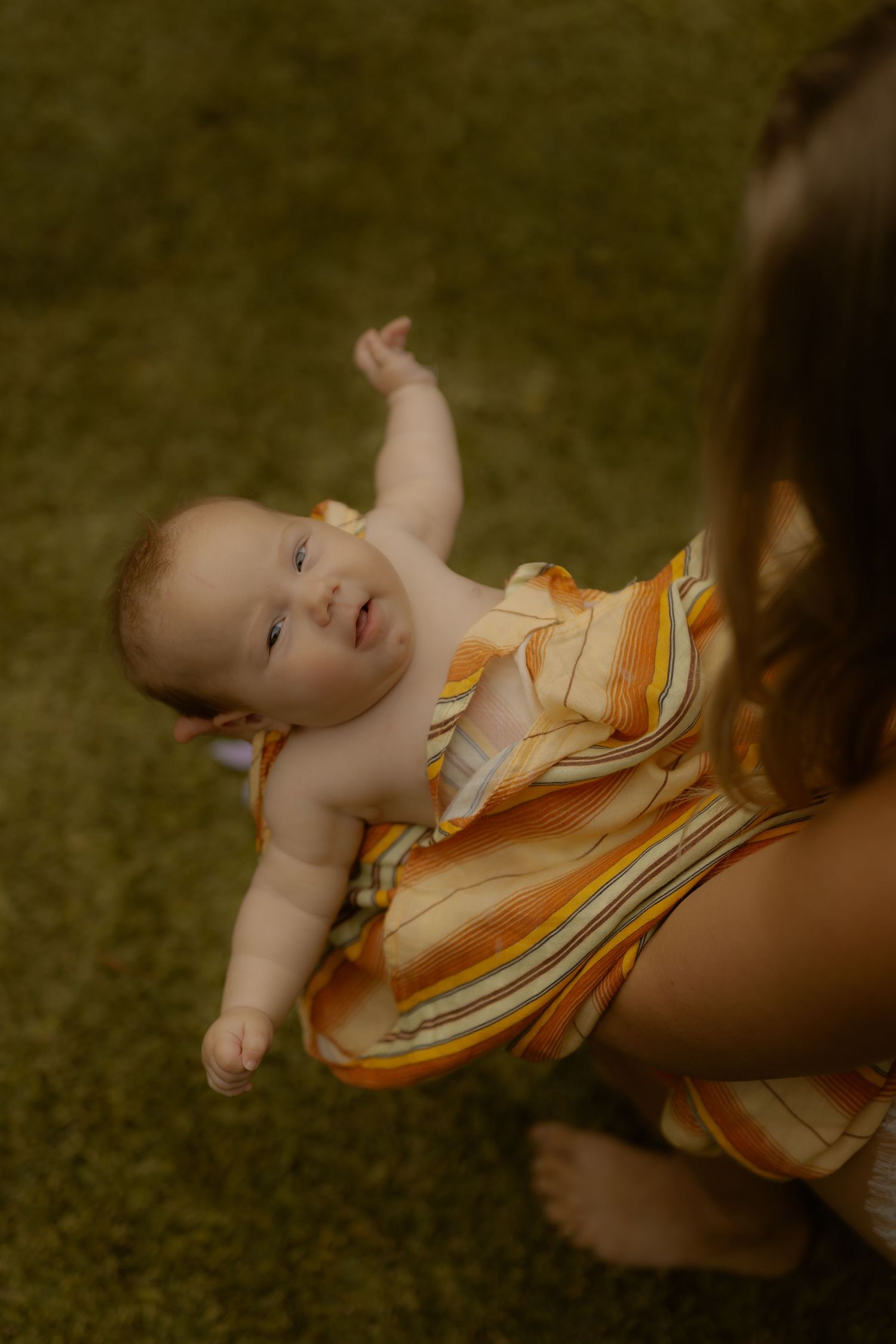 A baby in a striped orange romper being held up by a person kneeling on green carpet.