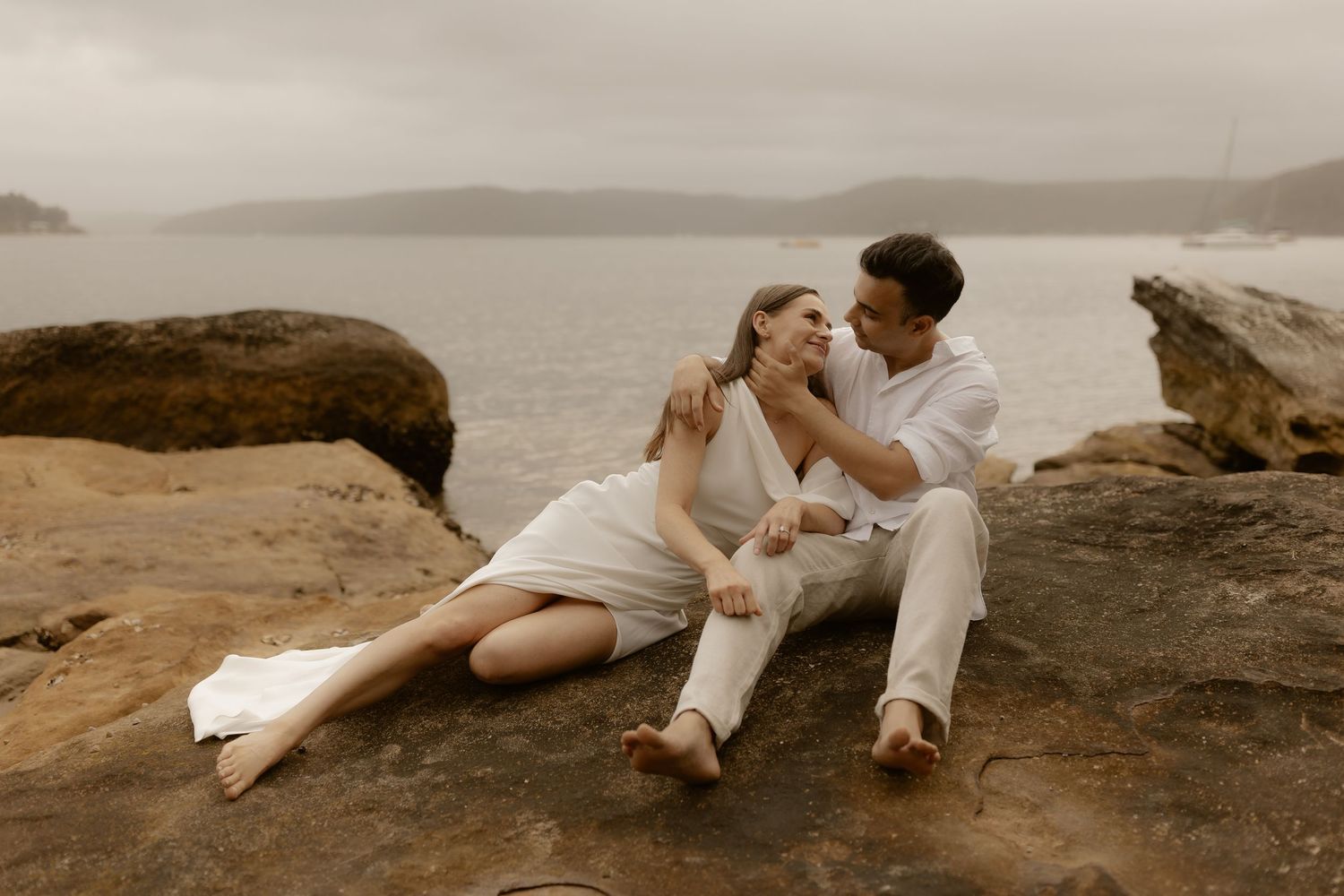 A couple shares romantic moments on coastal rocks overlooking the ocean during sunset.