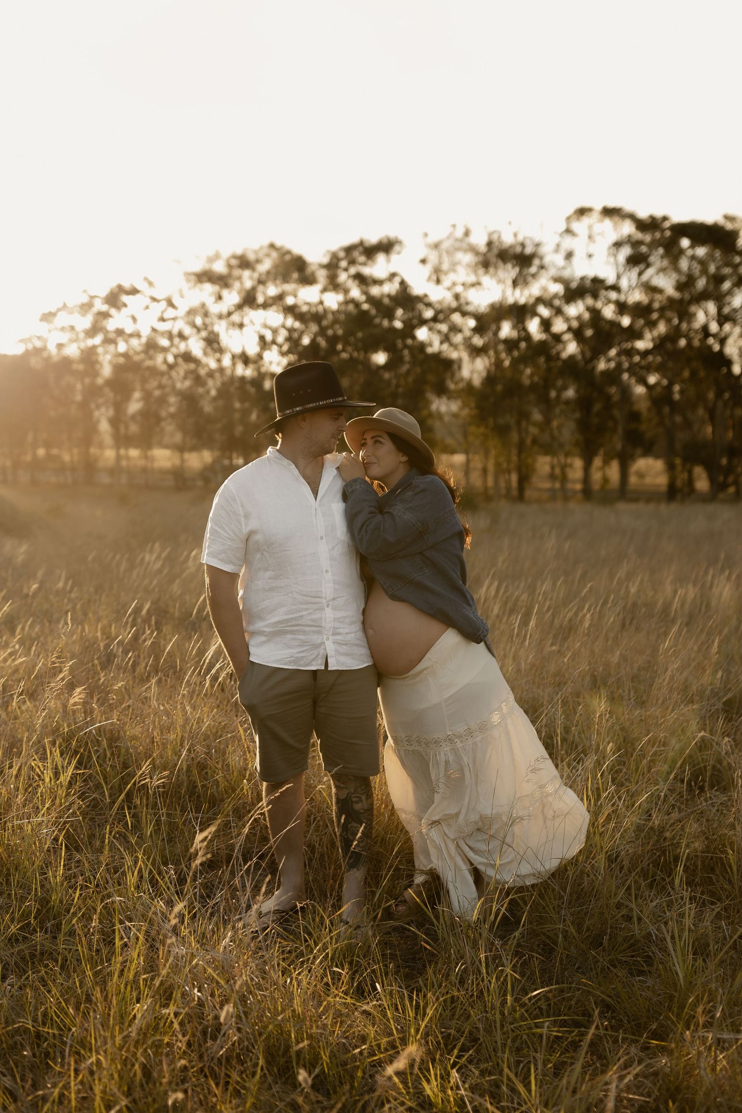 Couple embraces in a golden field at sunset with trees in background and warm sunlight filtering through.