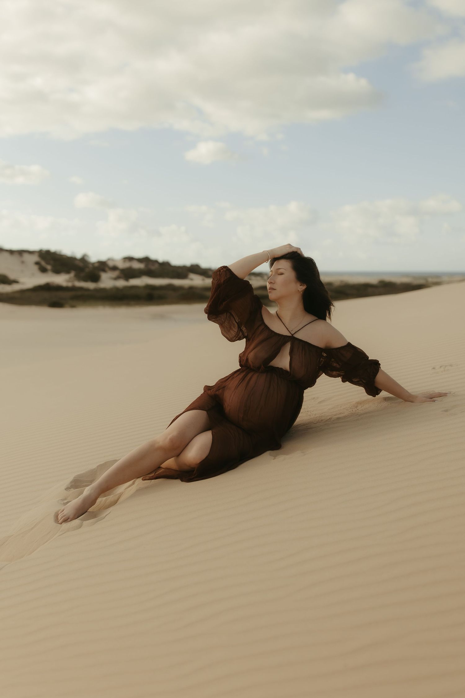 A person in dark attire reclines on the slope of a sandy desert dune beneath a partly cloudy sky.