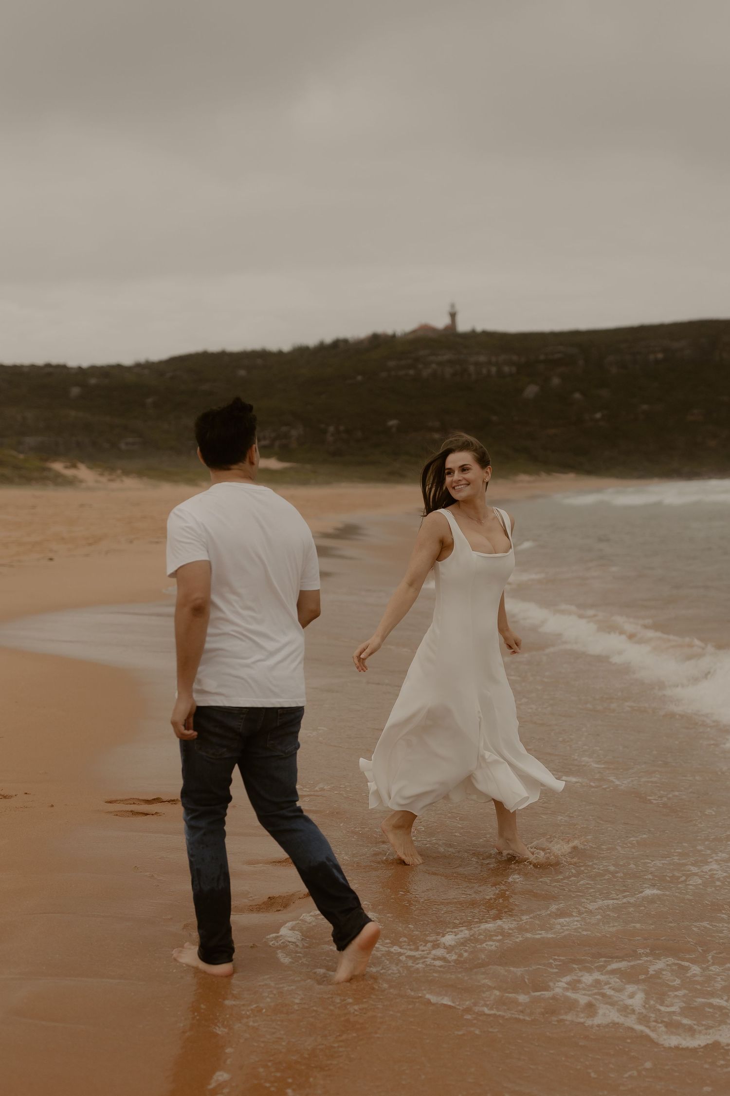A couple dances barefoot along the shoreline at sunset with rolling hills in the distance.