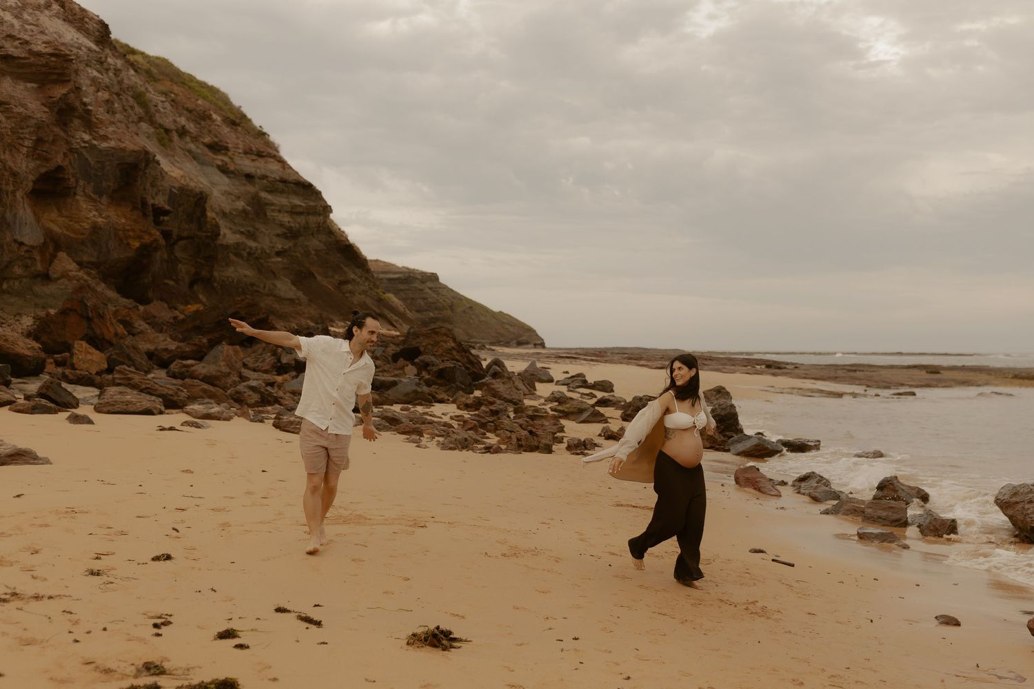 Couple walking and playing along a rocky beach shoreline during their wedding photo session.