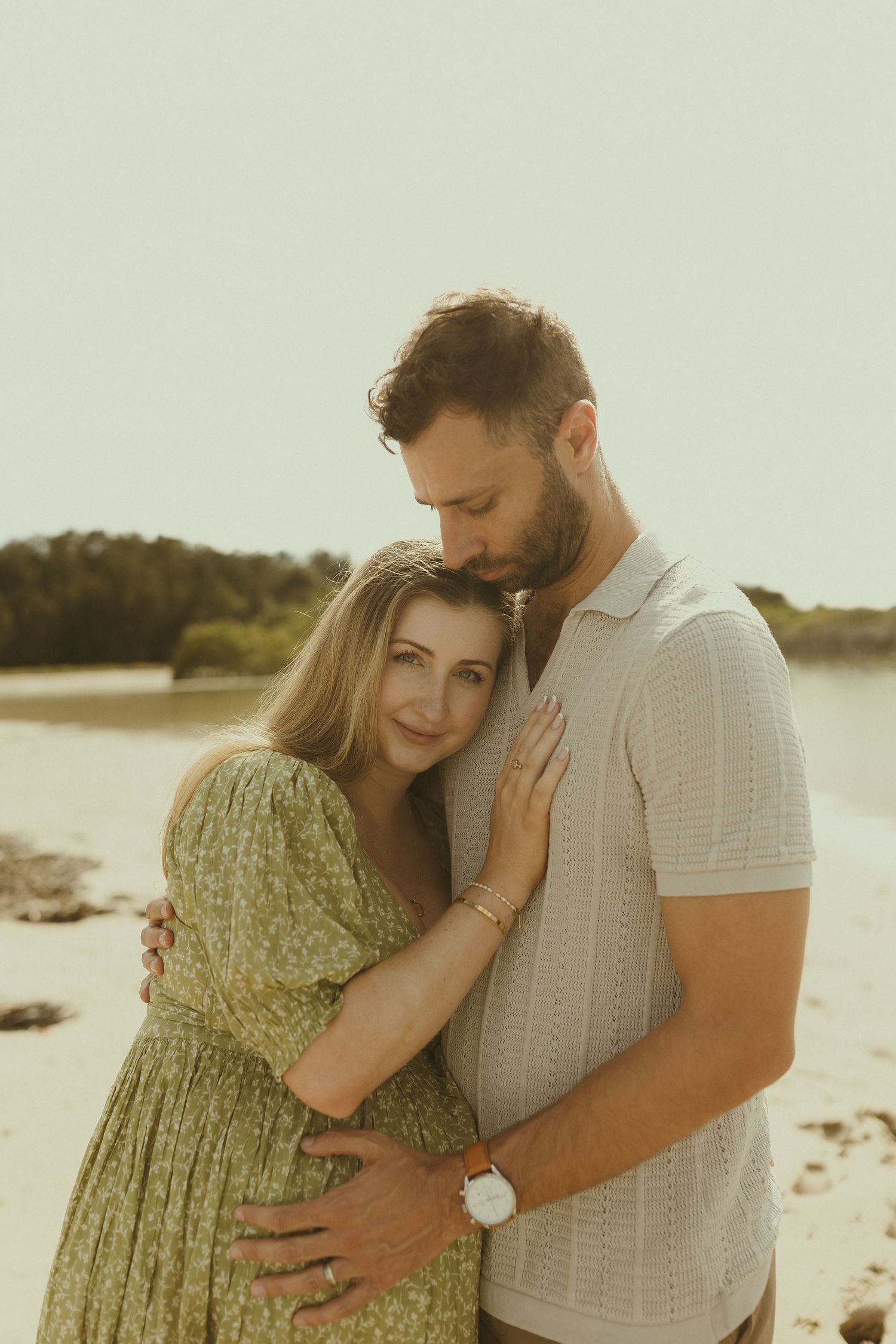 Couple embraces tenderly on a sandy beach during sunset wearing casual summer clothing.