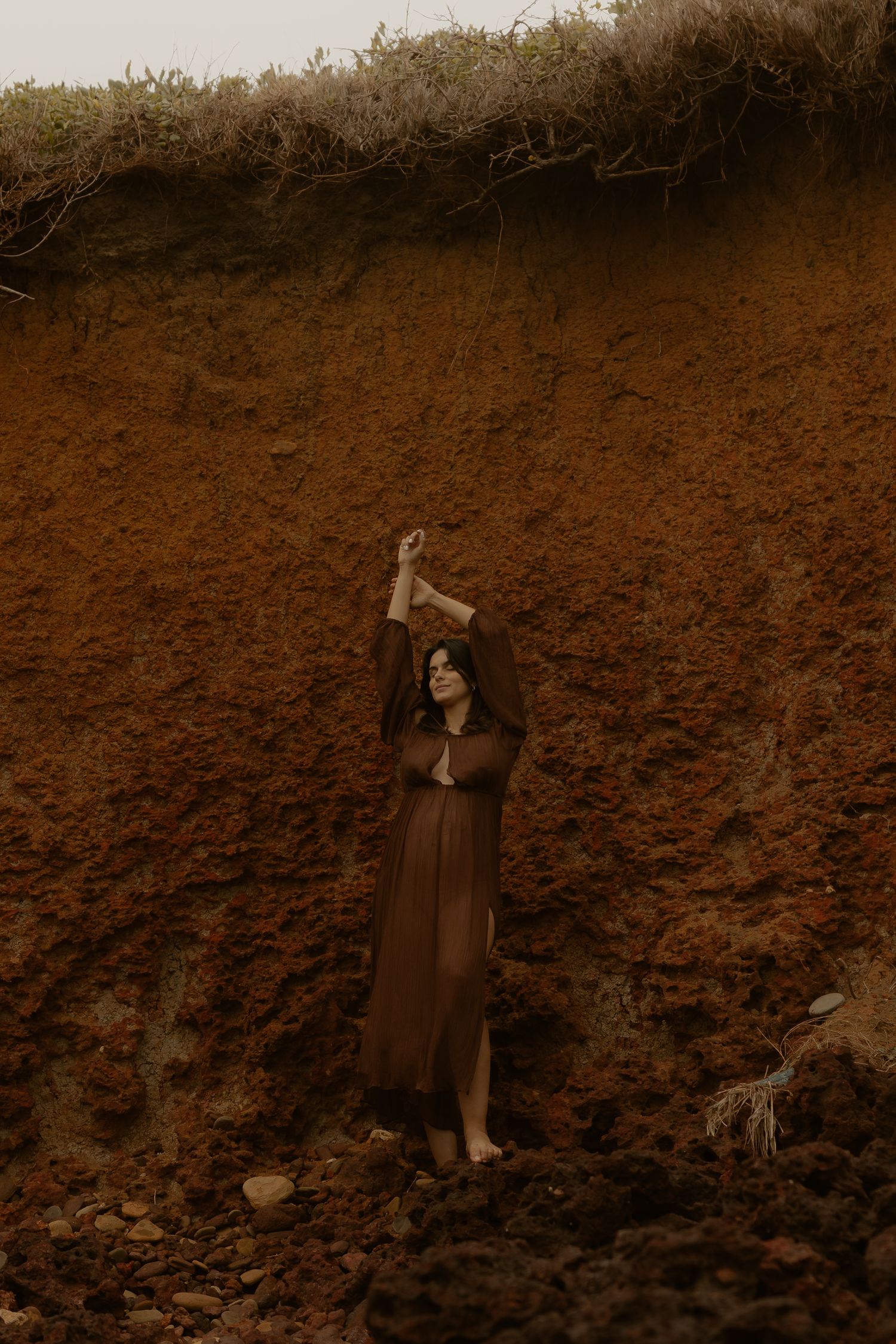 Expectant mother in a long brown dress poses against a rustic cliff wall during a maternity photoshoot.