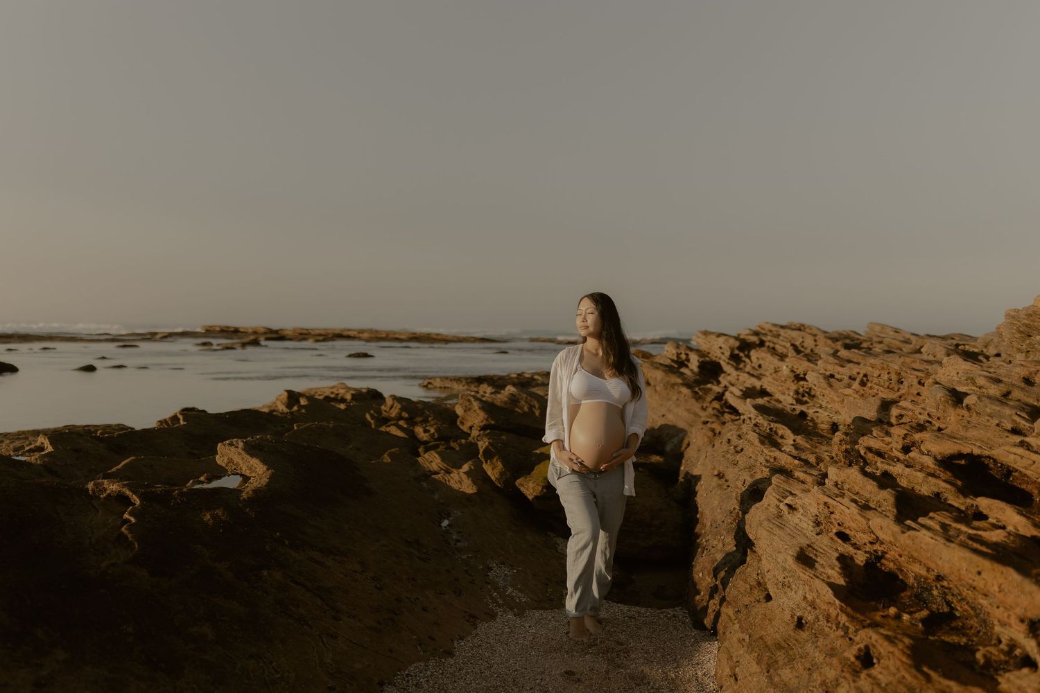 Person in white outfit walking along rocky coastal terrain with ocean views at sunset.