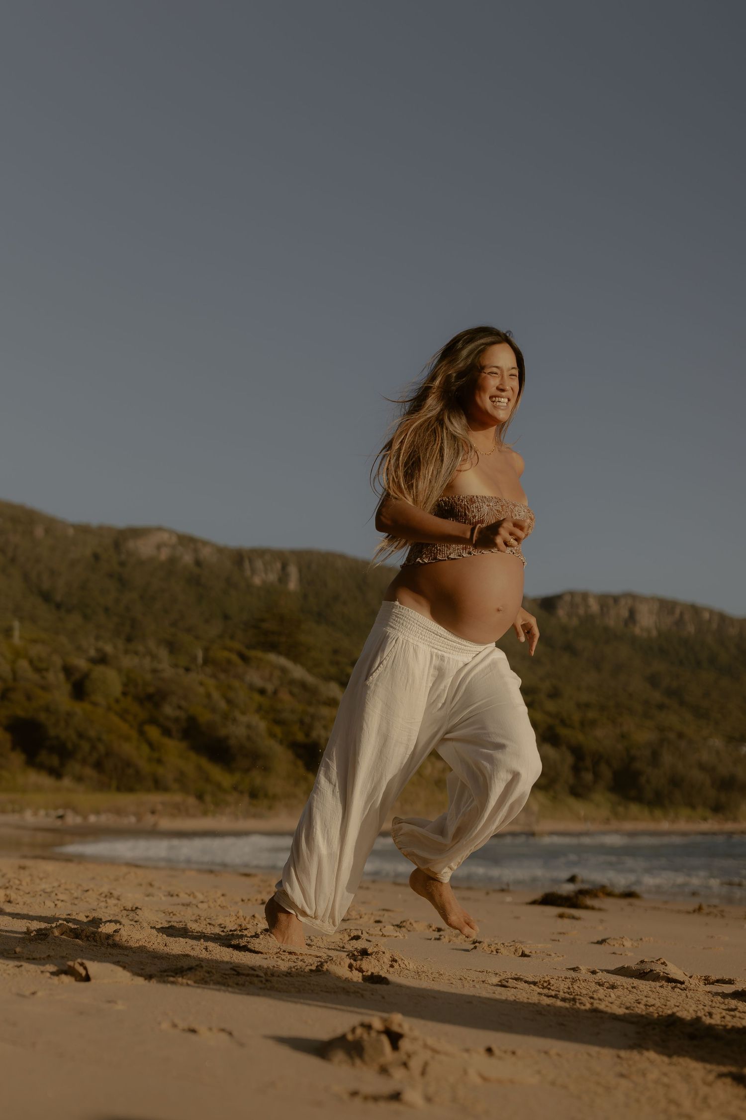 Expectant mother in white pants runs joyfully along a beach at sunset with mountains in the background.