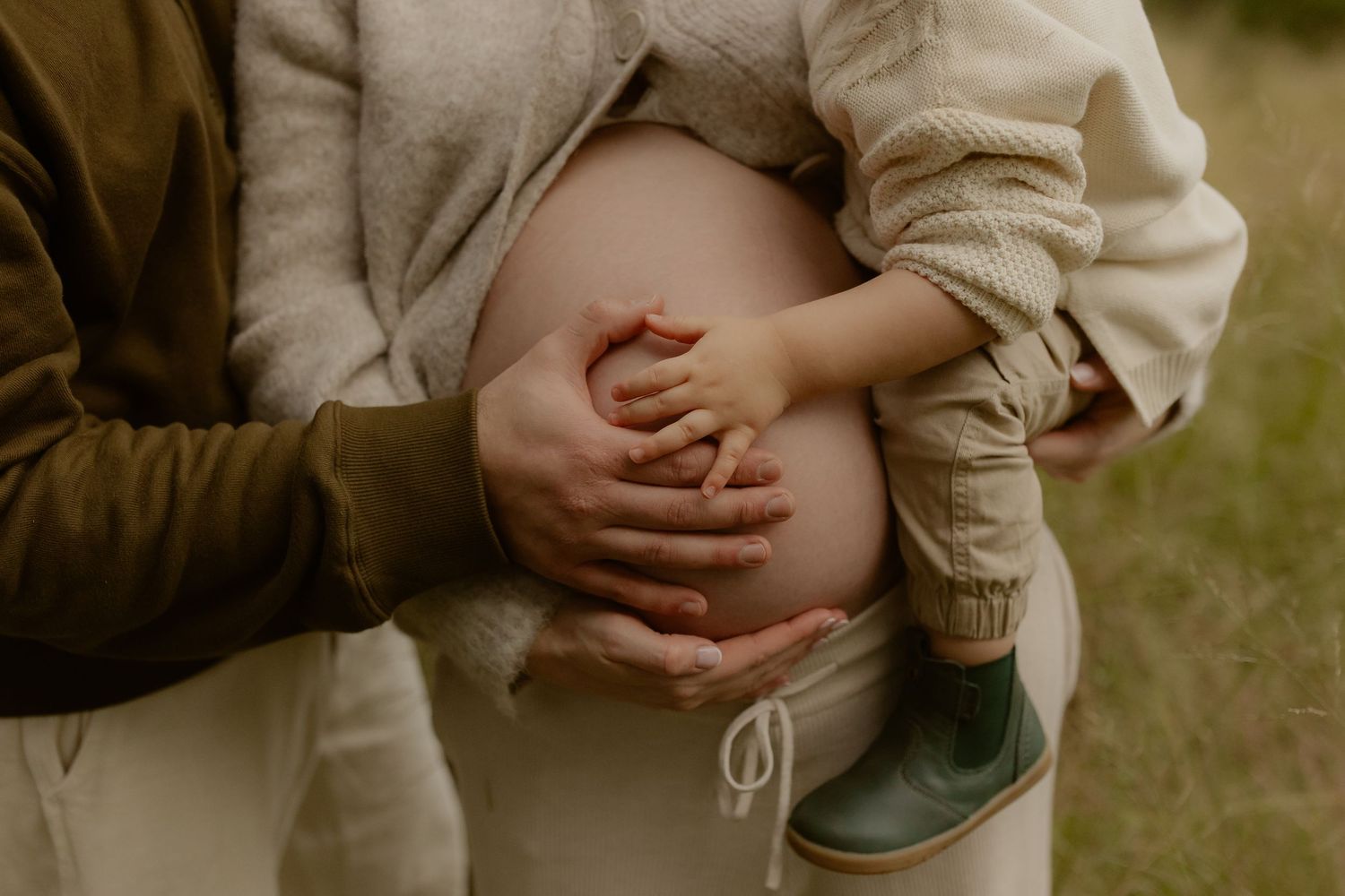 Pregnant woman's bare belly held tenderly by partner and toddler child outdoors in natural setting.