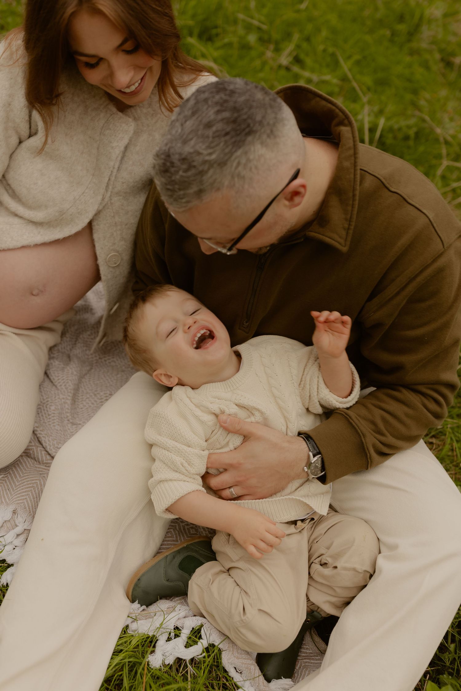 Parents laugh with their giggling toddler during a relaxed outdoor picnic on a green grassy hill.