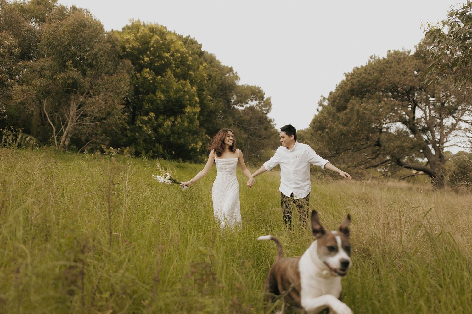 A couple in white outfits walks through a tall grass meadow during a sunny afternoon photo session.