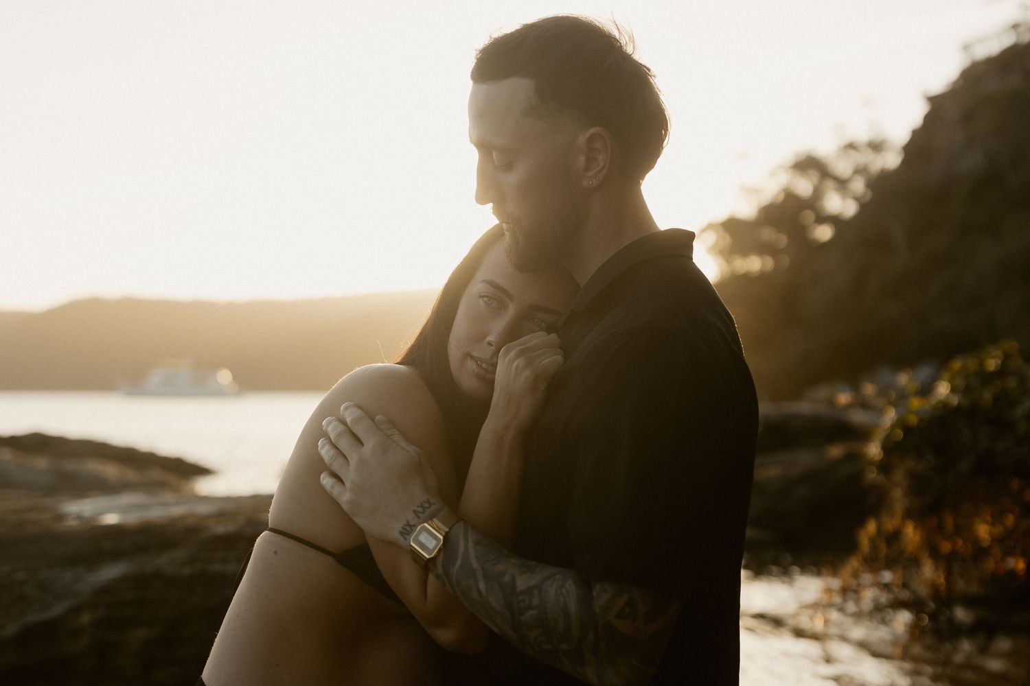 A couple embraces on a rocky coastline during golden hour as sunlight filters through misty ocean air.