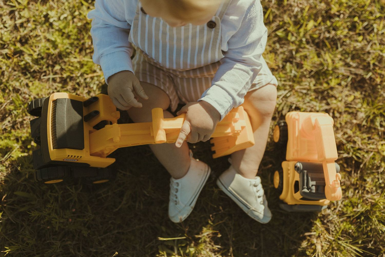 A baby in striped overalls plays with a wooden toy truck on sunny grass.