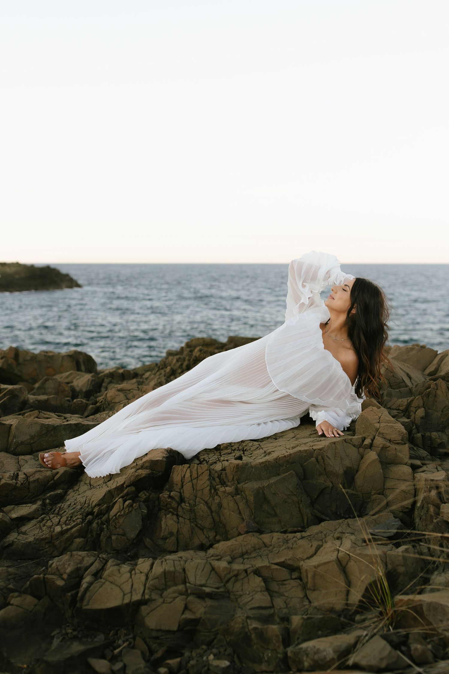 Woman in ethereal white dress reclines on dramatic rocky coastline.
