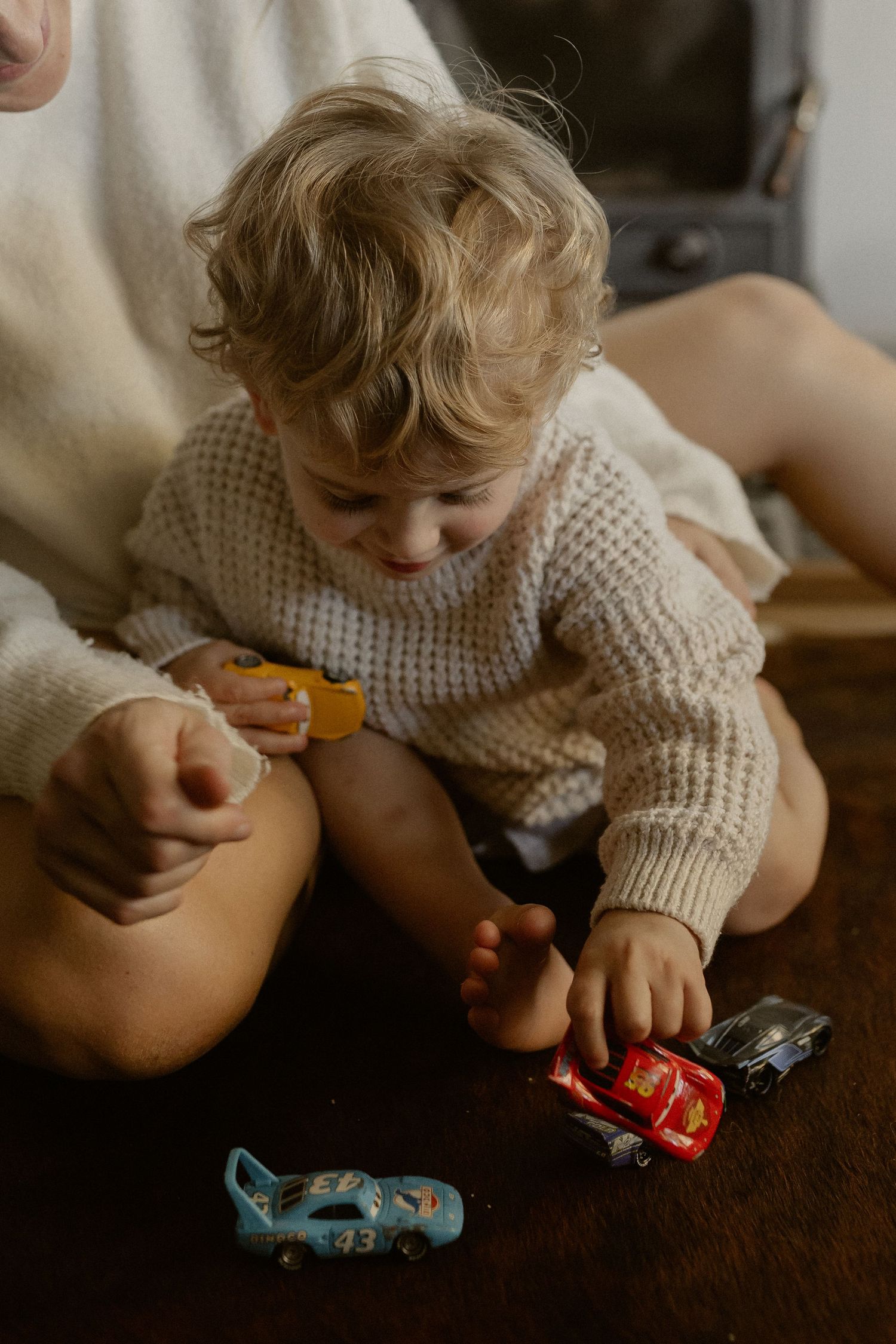 Young child in cream sweater plays with toy cars while sitting on dark surface.