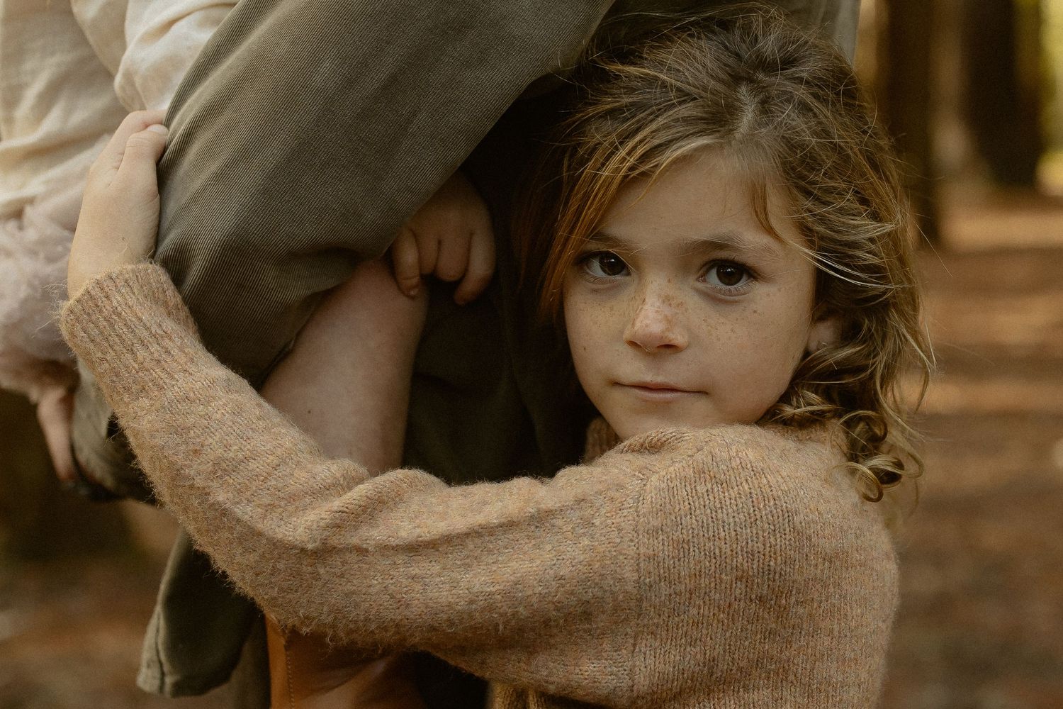 A child in a beige sweater gazes at the camera while holding onto someone's clothing in a warm-toned setting.