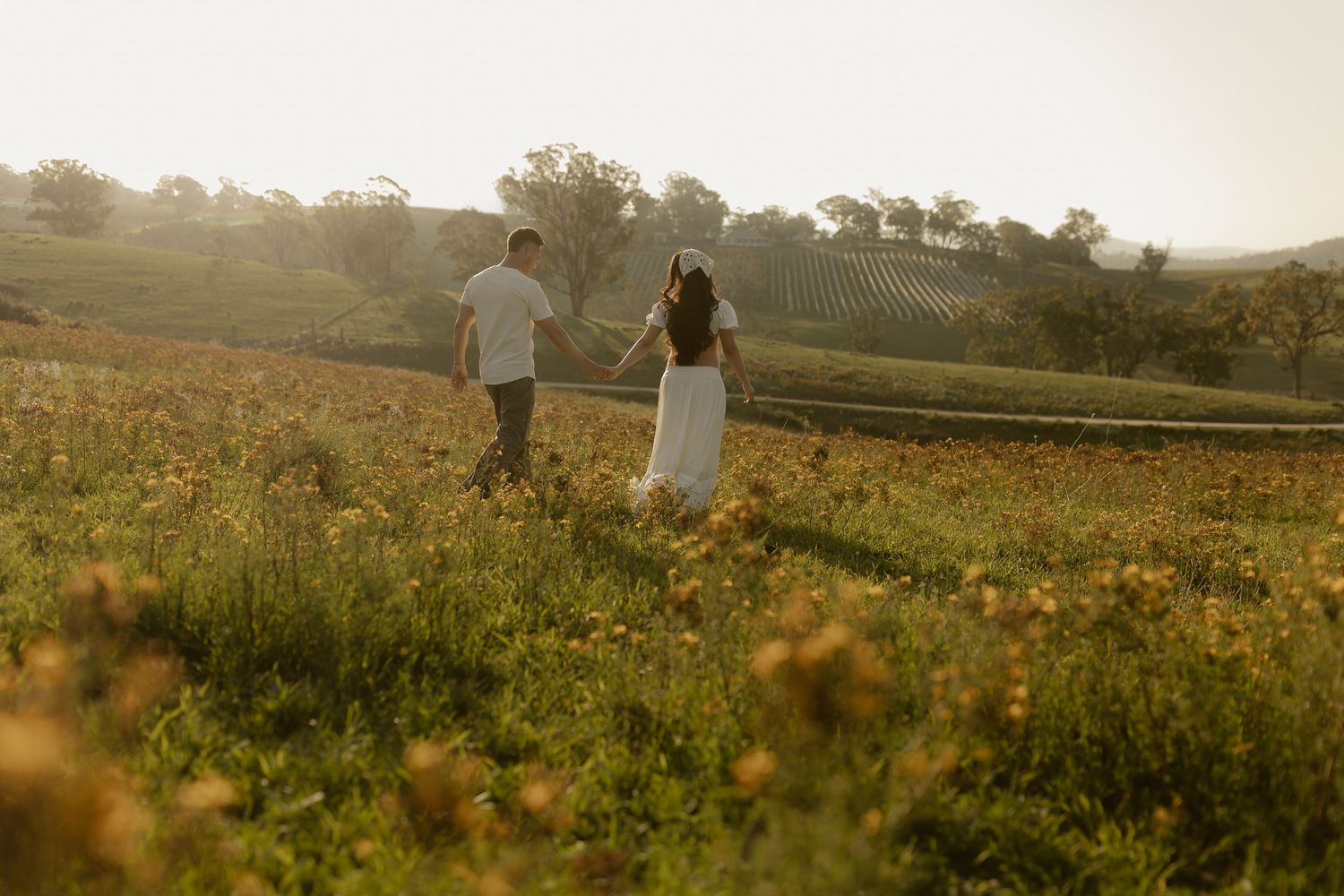 Silhouettes of two people walking through a golden meadow at sunset with rolling hills in the background.
