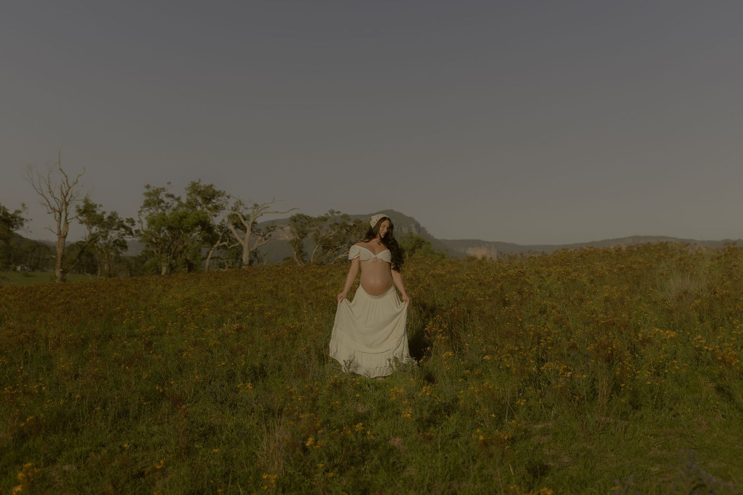 A couple embraces in a golden wildflower field during sunset with mountains in the background.