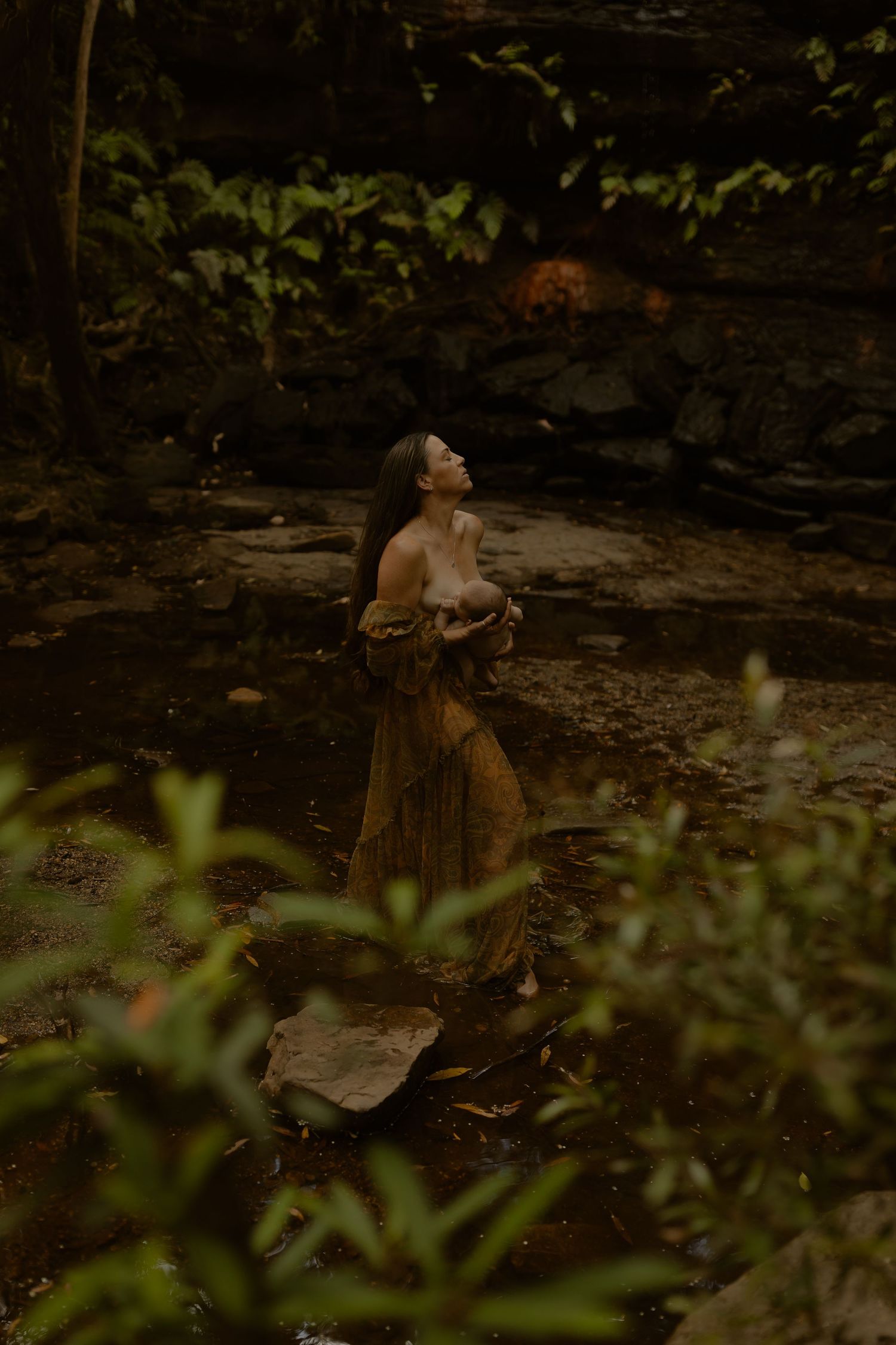 Atmospheric woodland scene with figures by a rocky stream in muted earth tones.