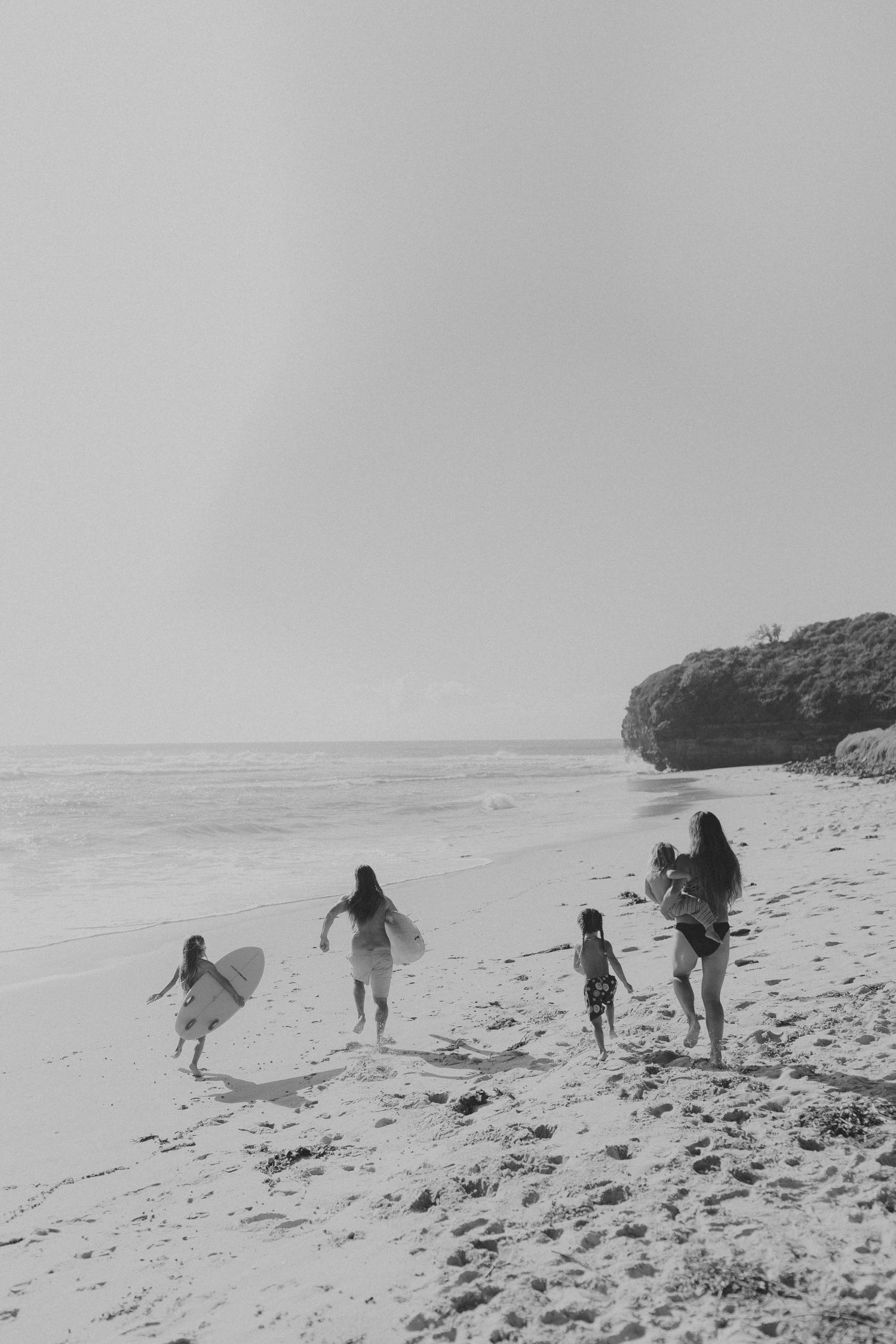 A group walking along a scenic beach carrying surfboards in black and white.