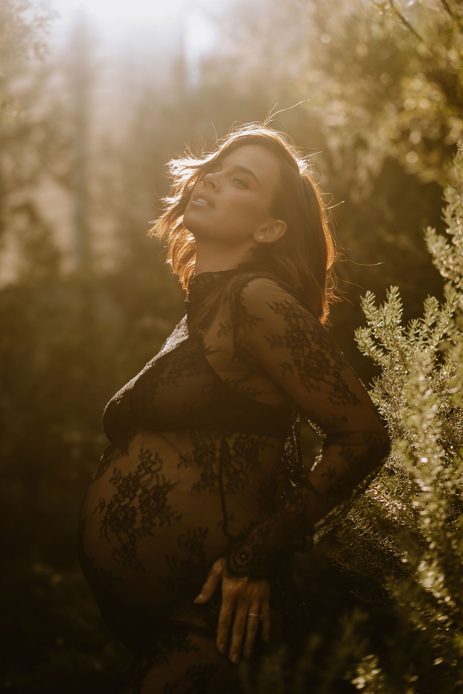 A pregnant woman in a black lace dress poses in golden sunset light amongst foliage during a maternity photoshoot.