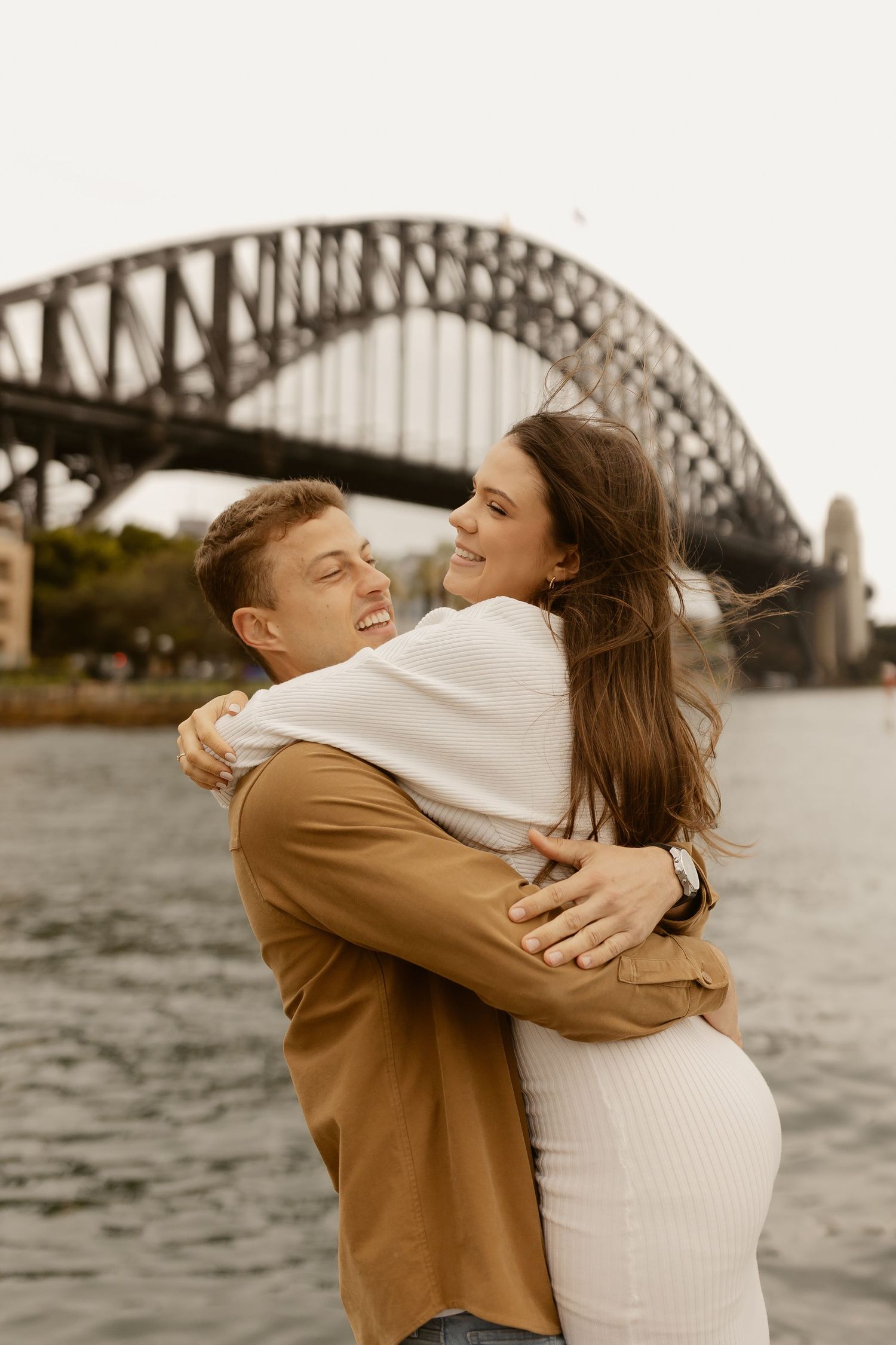Couple embracing passionately in front of Sydney Harbour Bridge at sunset.