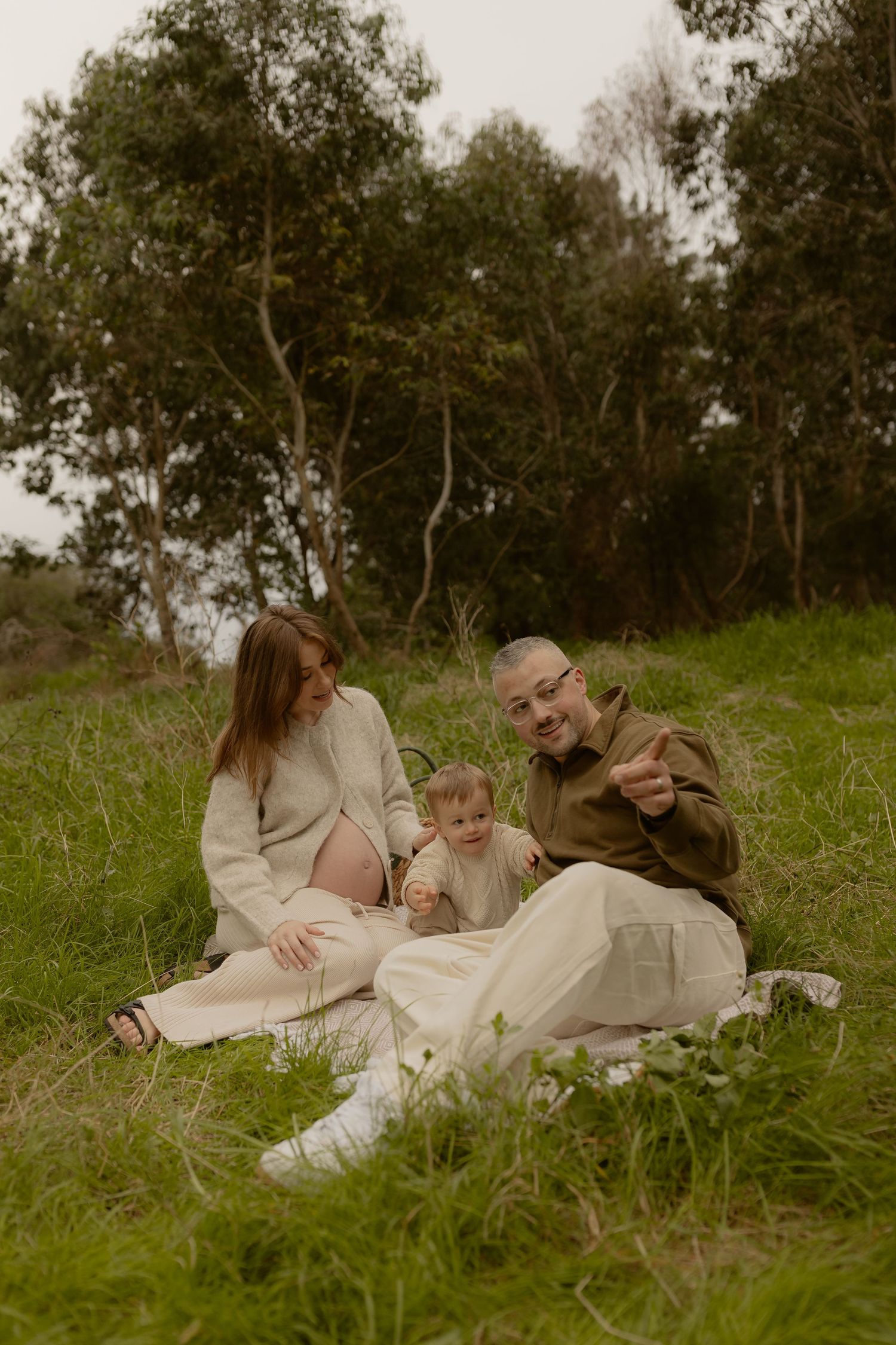 A happy family with a baby sits on grass in a park, pregnant mother in white beside father in olive shirt.