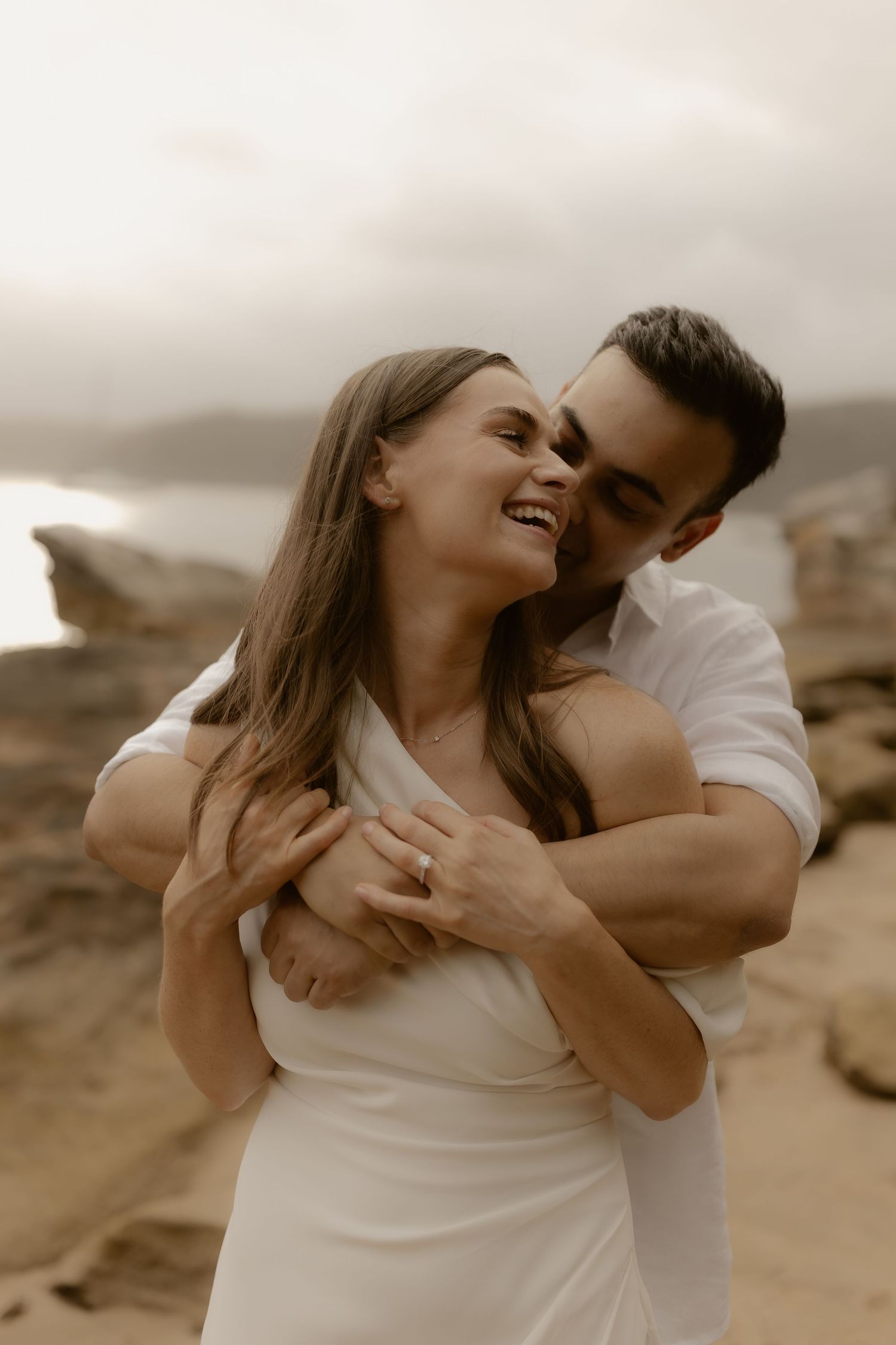 A couple shares an intimate moment together on a scenic beach during a romantic photoshoot in sepia tones.