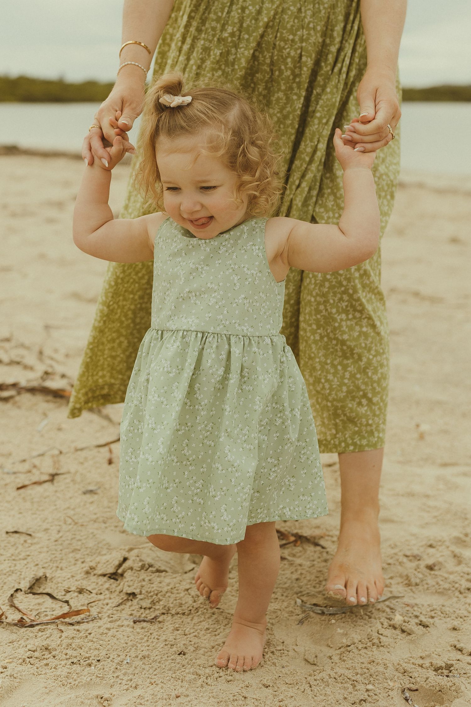 Small bare feet stand in the sand as a child in a light dress plays at the beach during golden hour.