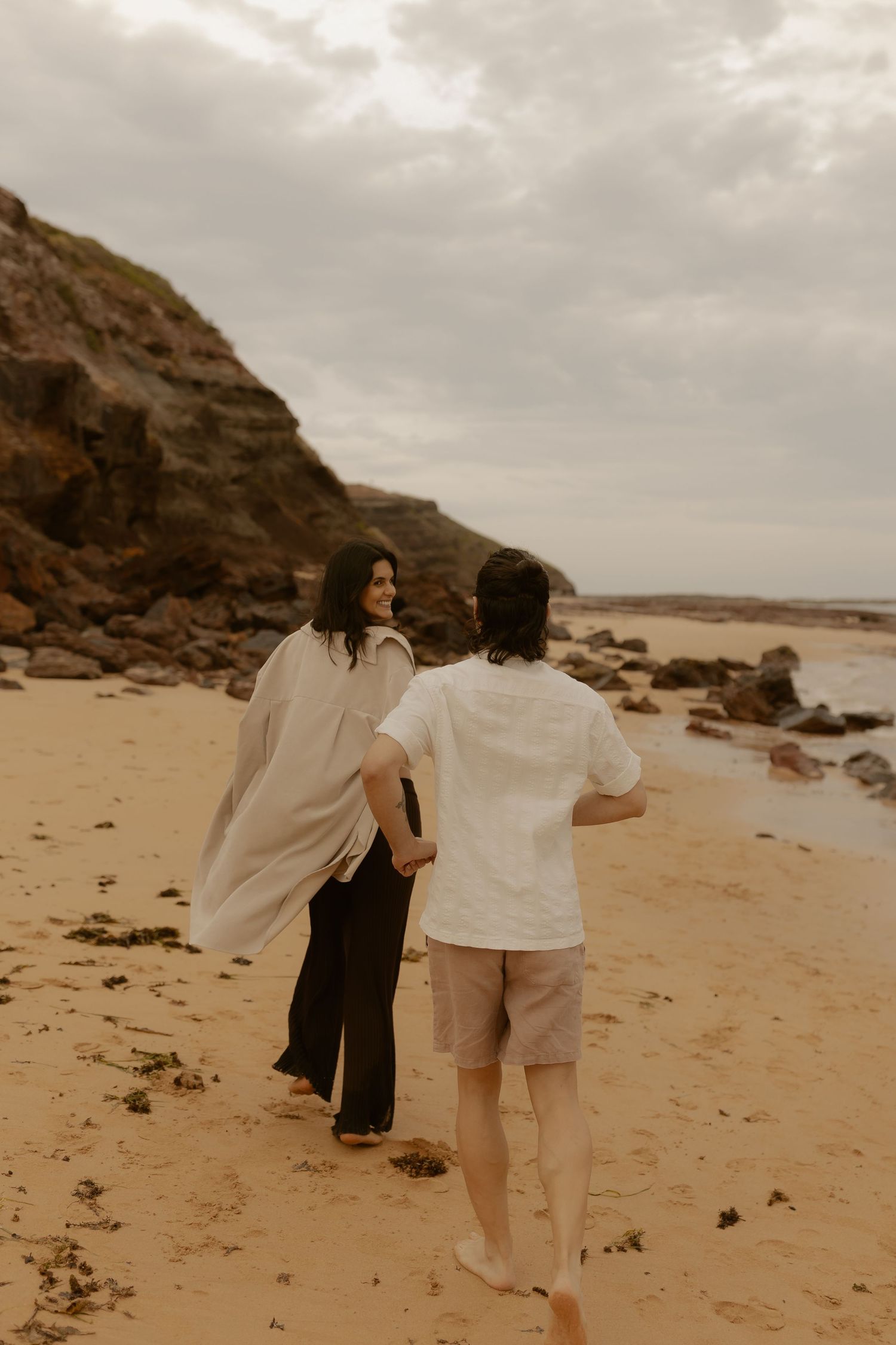 A couple walks hand in hand along a scenic beach with rocky cliffs in the background.